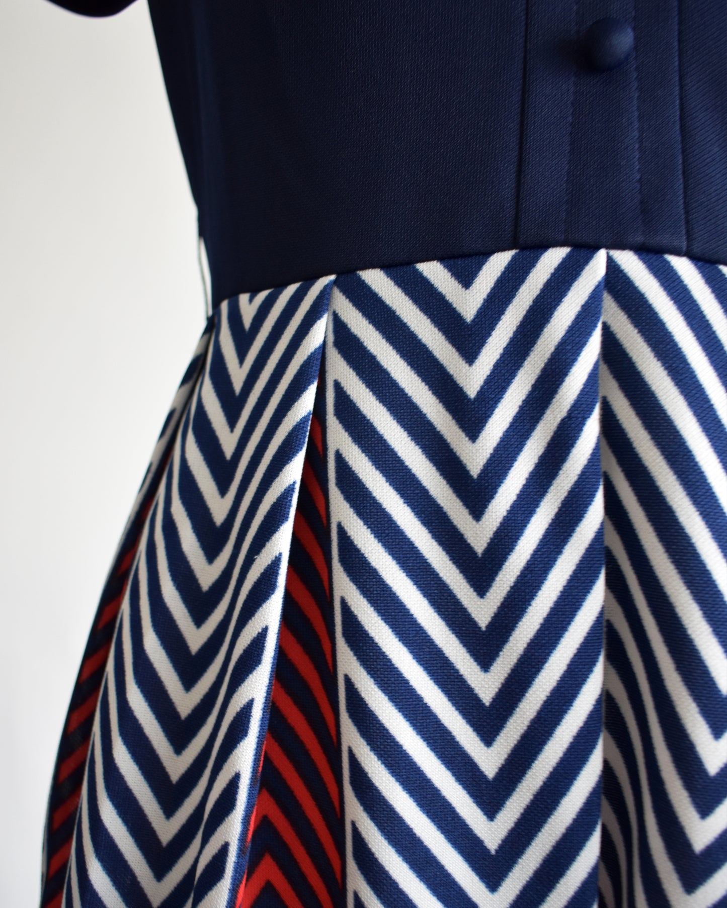 close up of the waist of a vintage 1970s navy blue, white, and red dress with a chevron patterned skirt on a mannequin against a white background