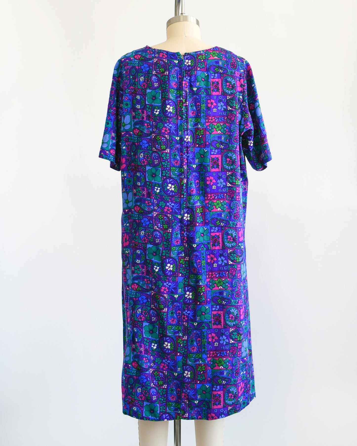 Back view of a vintage floral dress that has a vibrant purple, blue, pink, teal, green, and white psychedelic floral print. The dress has short sleeves, scoop neckline, zipper up the back, and is modeled on a dress form.