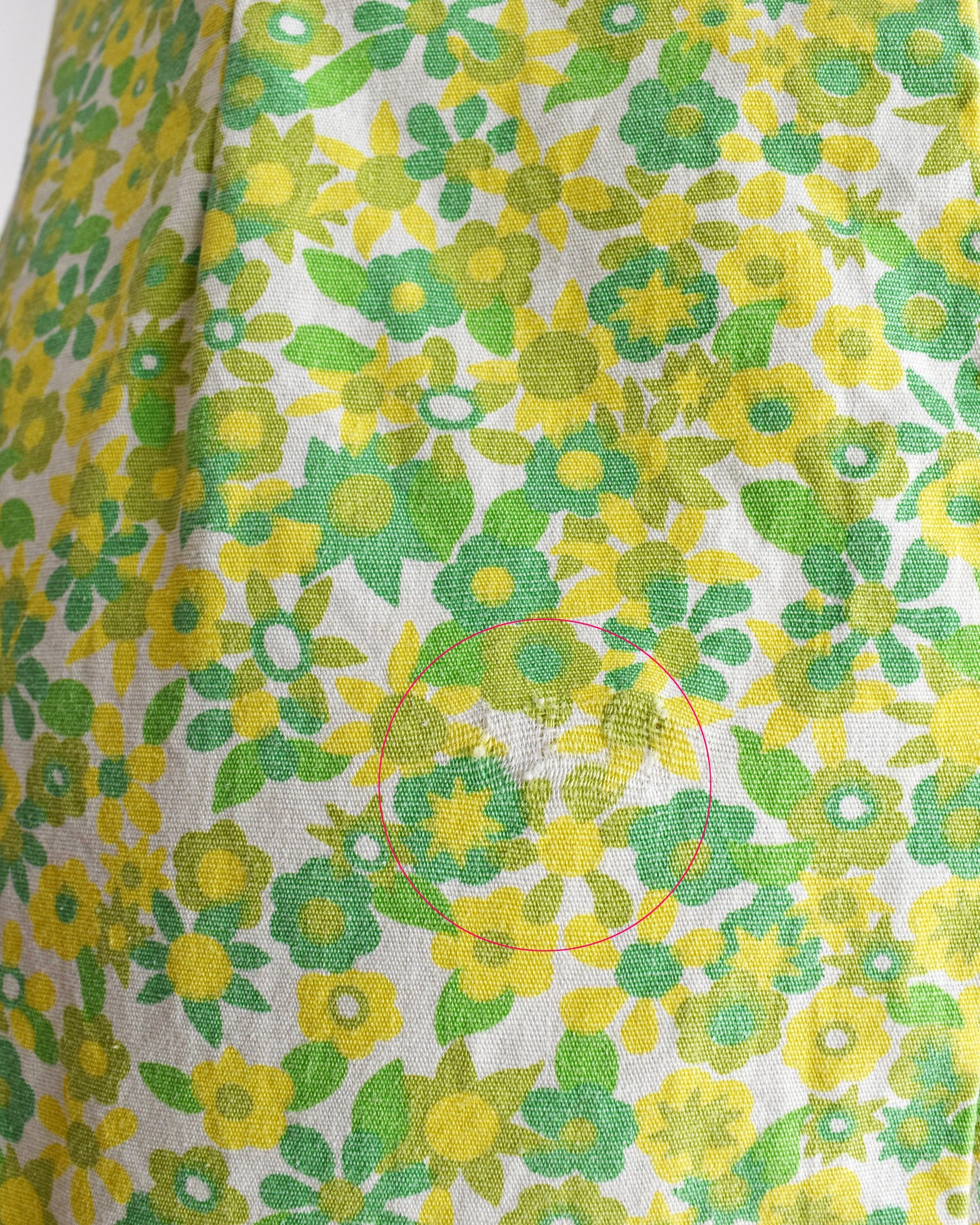A close-up of a the stressed fabric spot on a vintage 1960s sleeveless, A-line mini dress with a green and yellow floral pattern, displayed on a beige mannequin against a plain white background.