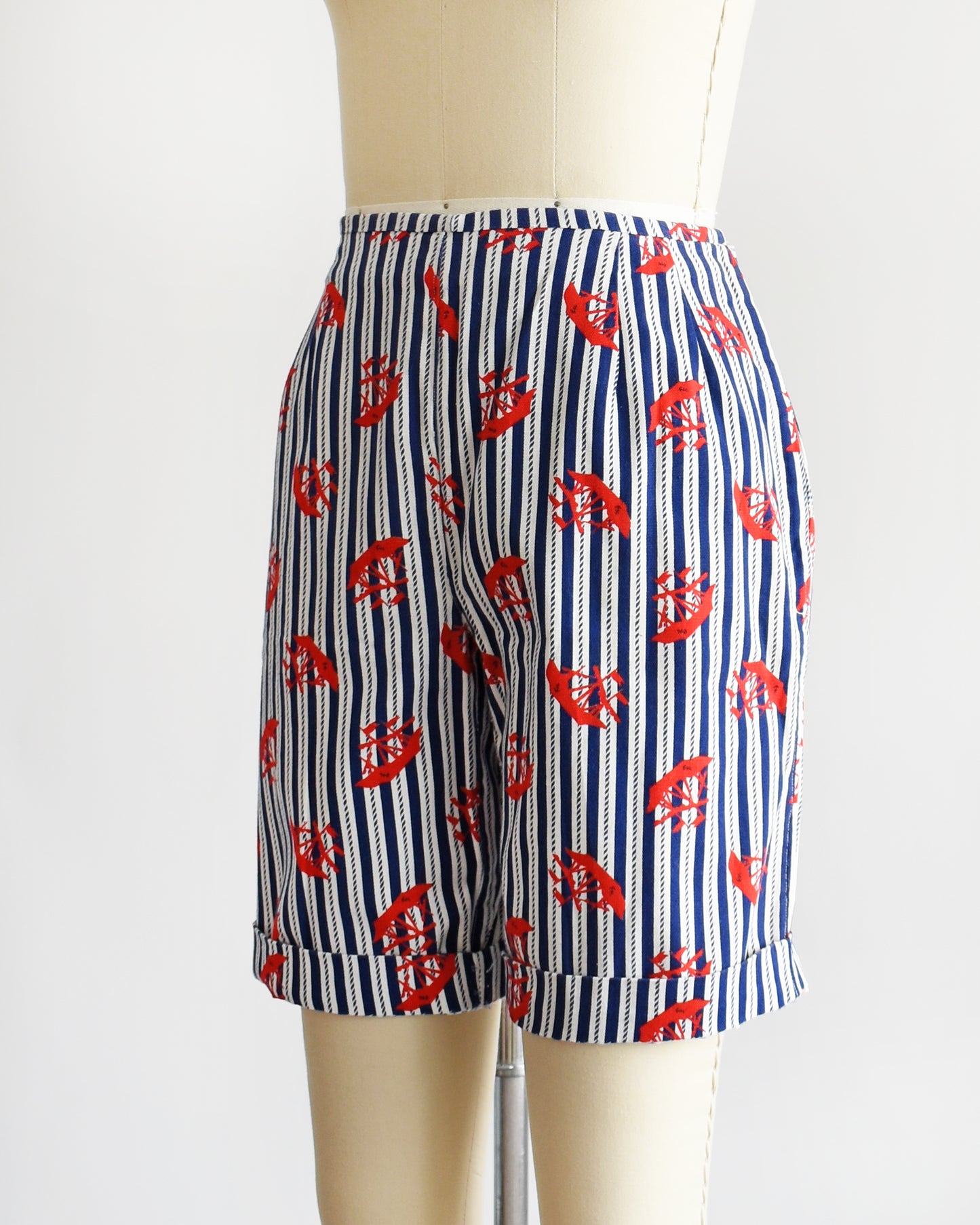 Side front view of a pair of vintage 60s shorts that are navy blue and white vertical striped with a red clipper ship print. The shorts are modeled on a dress form. The shorts are cuffed.