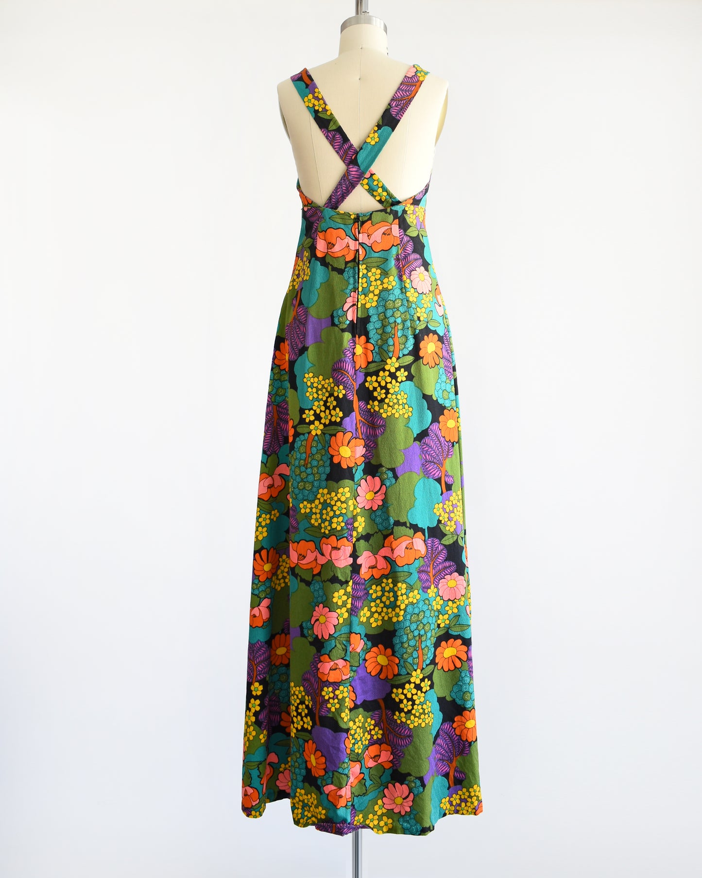 Back view of a vintage 70s floral maxi dress that is black and has a vibrant flower power print in greens, blue, peach, orange, yellow, purple, and white. Criss cross straps on the back. Dress is on dress form.