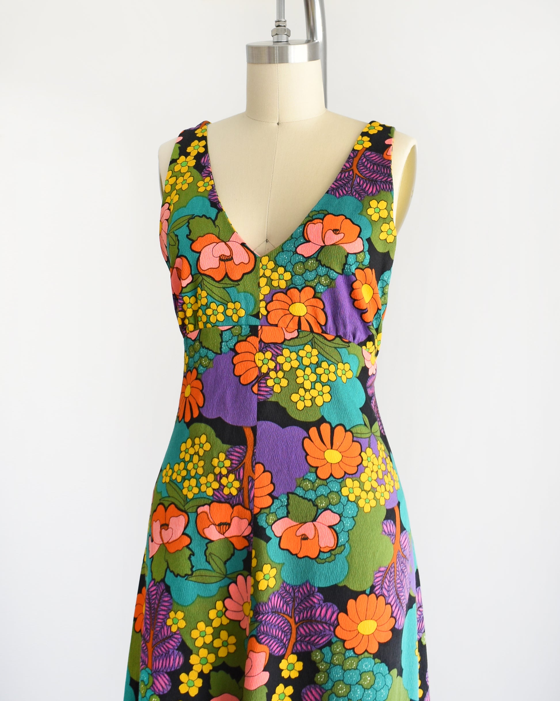 Side front view of a vintage 70s floral maxi dress that is black and has a vibrant flower power print in greens, blue, peach, orange, yellow, purple, and white. V-neckline. Dress is on dress form.