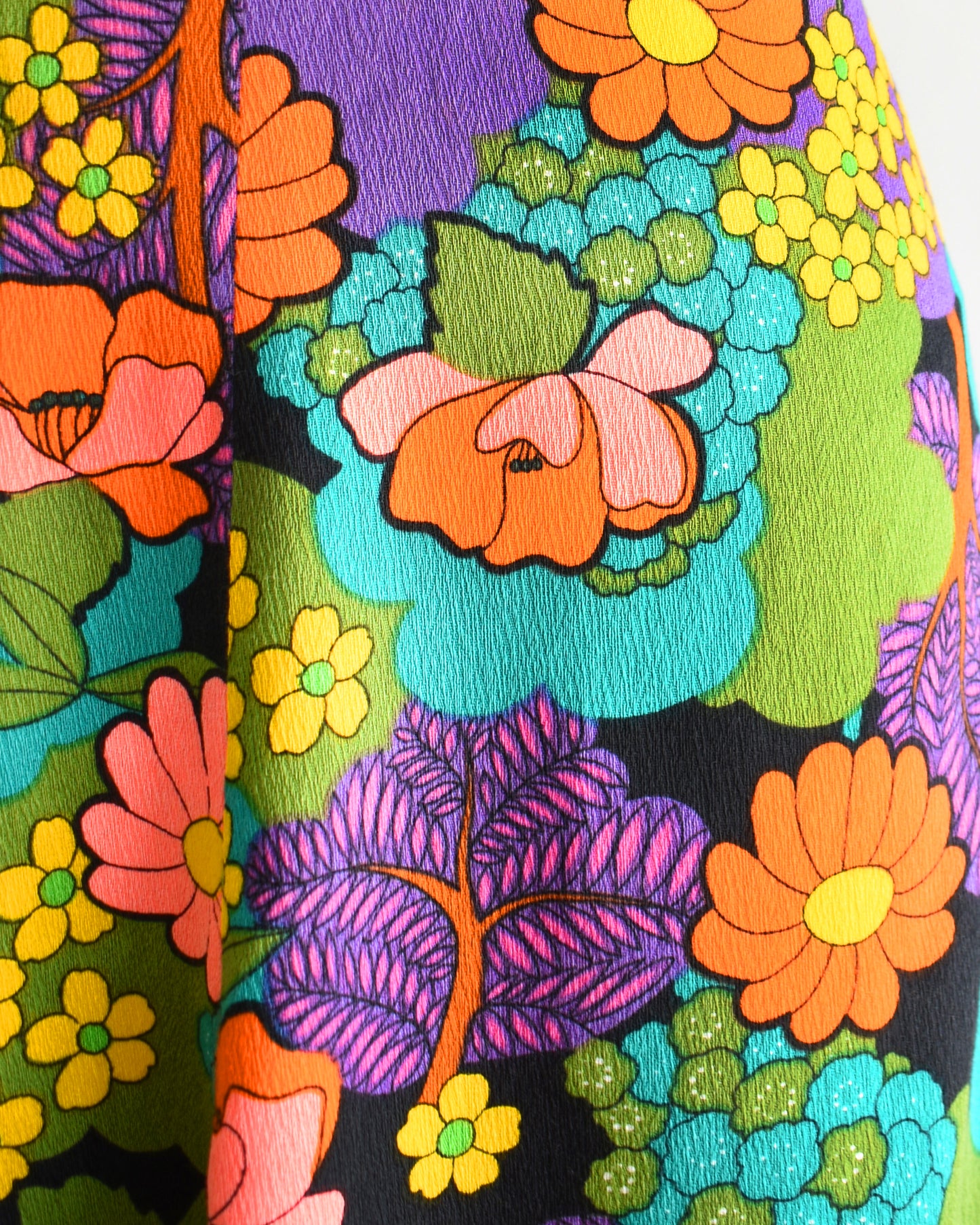 Close up of the brant flower power print in greens, blue, peach, orange, yellow, purple, and white set on black. V-neckline. Dress is on dress form.