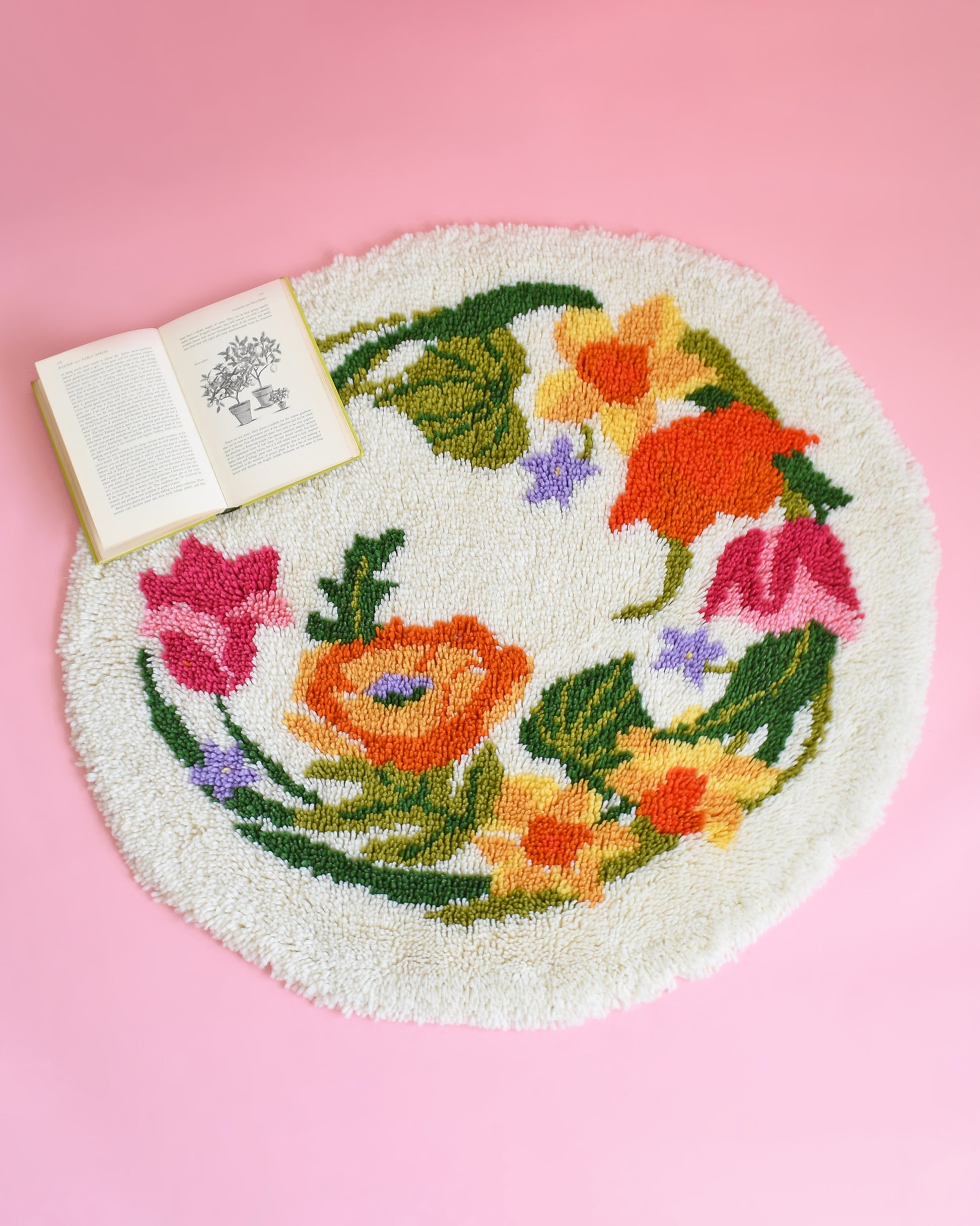 A vintage round floral latch hook rug that has cream white shaggy acrylic with a colorful floral motif in pinks, orange, yellow, purple and greens. The rug is on a pink background and has an open book on it for scale.