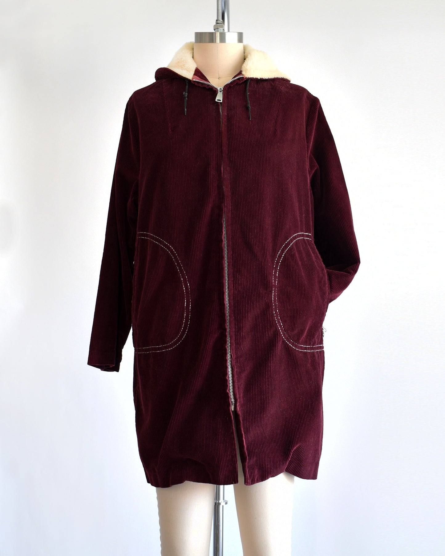 a vintage 1970s burgundy corduroy jacket with a sherpa lined hood on a dress form. One of the arms is tucked into a pocket.
