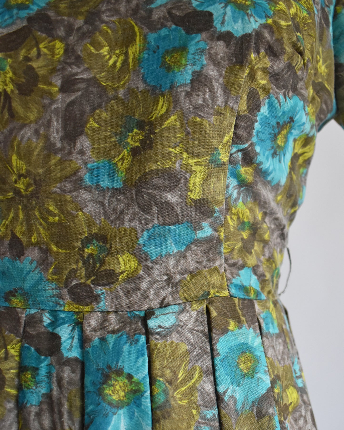 Close-up of a floral dress fabric with large yellow-green and turquoise flowers on a gray background, showing the pleated waistband detail.