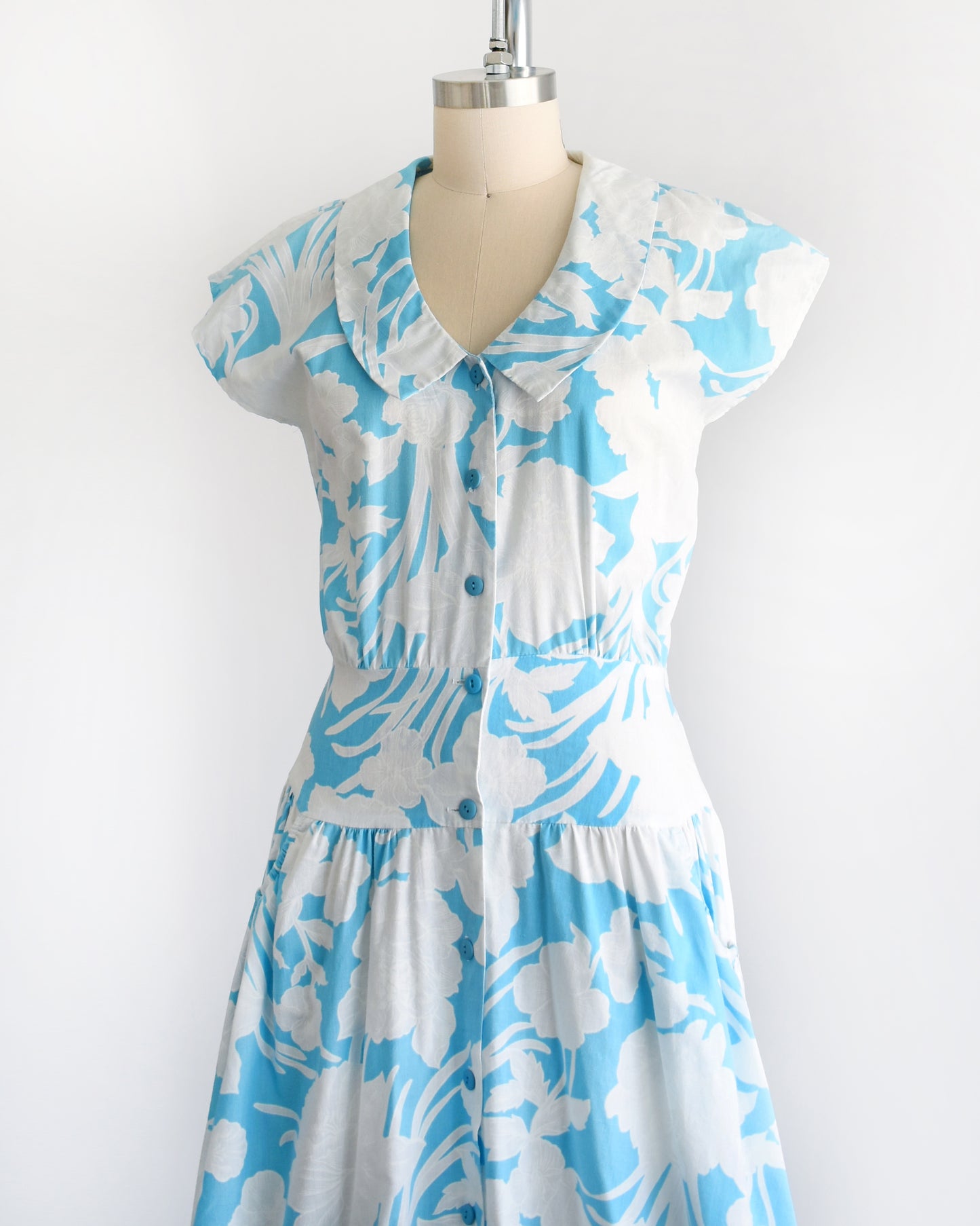 Side front view of a vintage 80s floral dress that is light blue with white floral print. Peter Pan style collar with blue buttons down the front. Cap sleeves. Wide waistband and drop waist skirt.