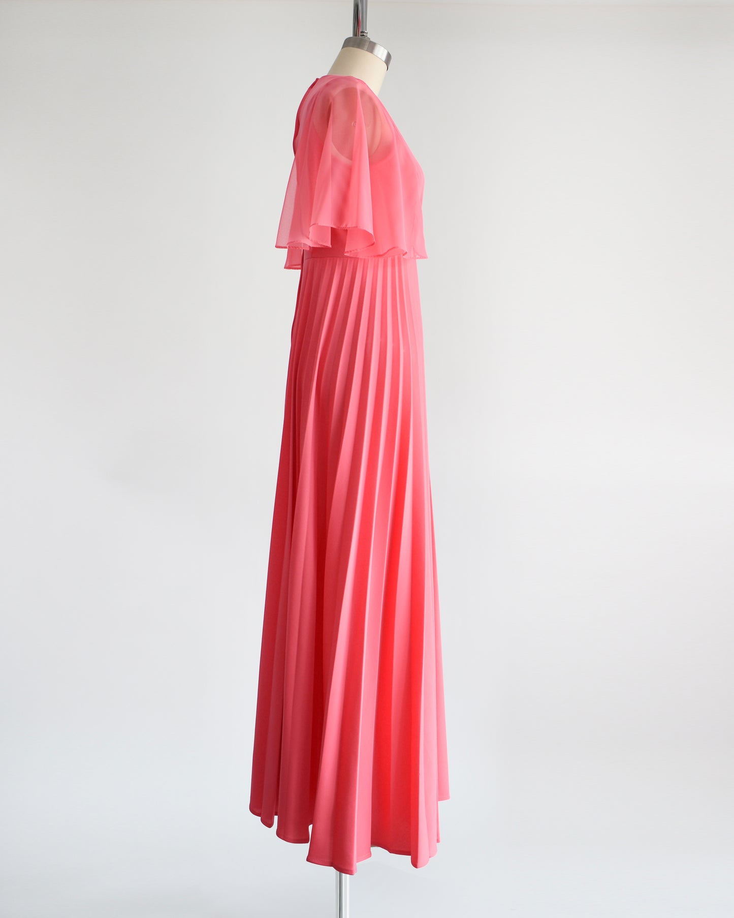 Side view of a vintage 70s coral pink maxi dress that has a faux wrap front with semi-sheer flutter sleeves, empire waist, and a long flowing accordion pleated skirt. The dress is on a dress form.