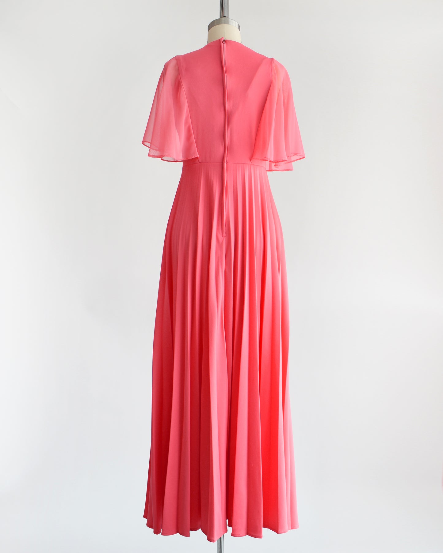 Back view of a vintage 70s coral pink maxi dress that has a faux wrap front with semi-sheer flutter sleeves, empire waist, and a long flowing accordion pleated skirt. Zipper up the back. The dress is on a dress form.