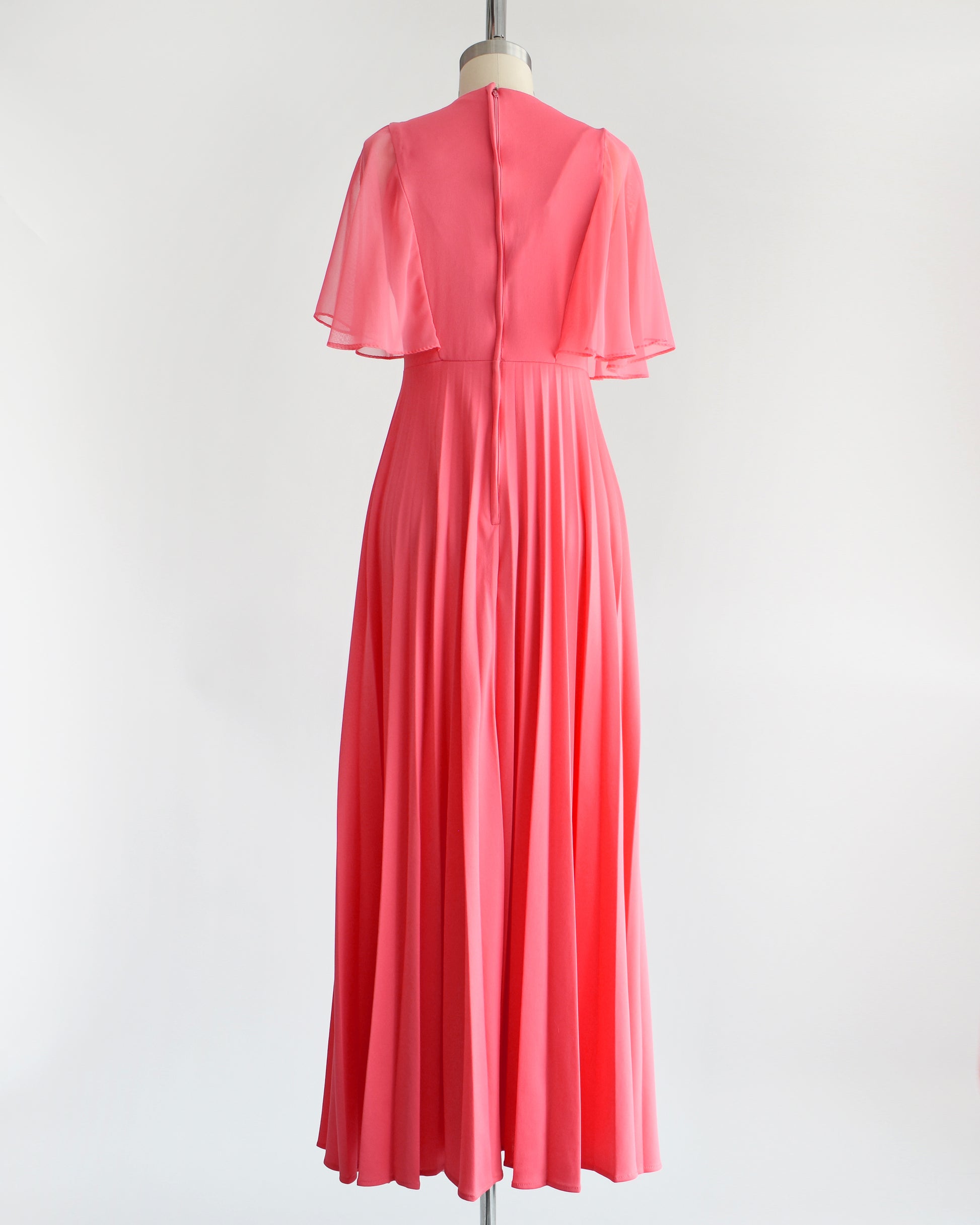 Back view of a vintage 70s coral pink maxi dress that has a faux wrap front with semi-sheer flutter sleeves, empire waist, and a long flowing accordion pleated skirt. Zipper up the back. The dress is on a dress form.