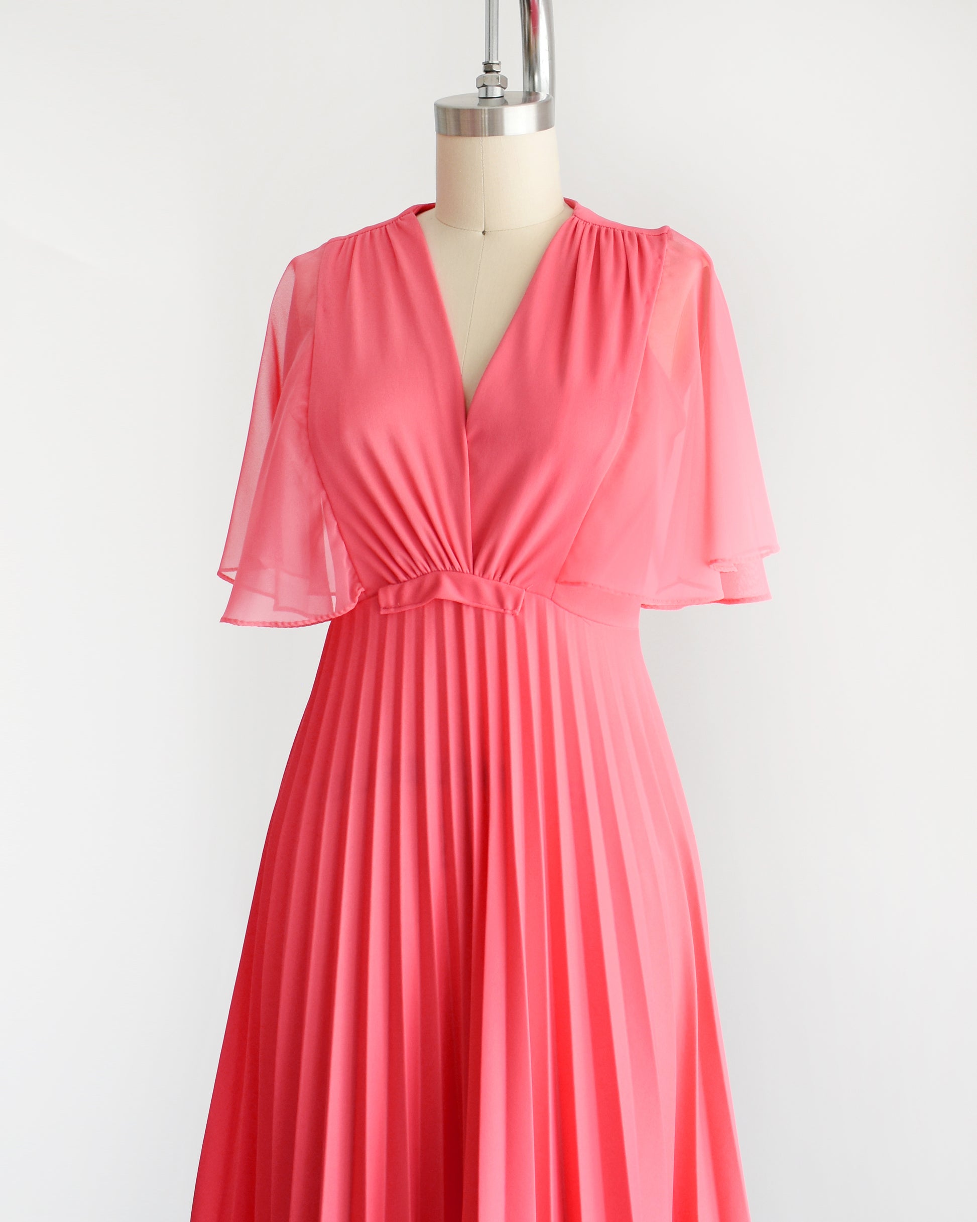 Side front view of a vintage 70s coral pink maxi dress that has a faux wrap front with semi-sheer flutter sleeves, empire waist, and a long flowing accordion pleated skirt. The dress is on a dress form.