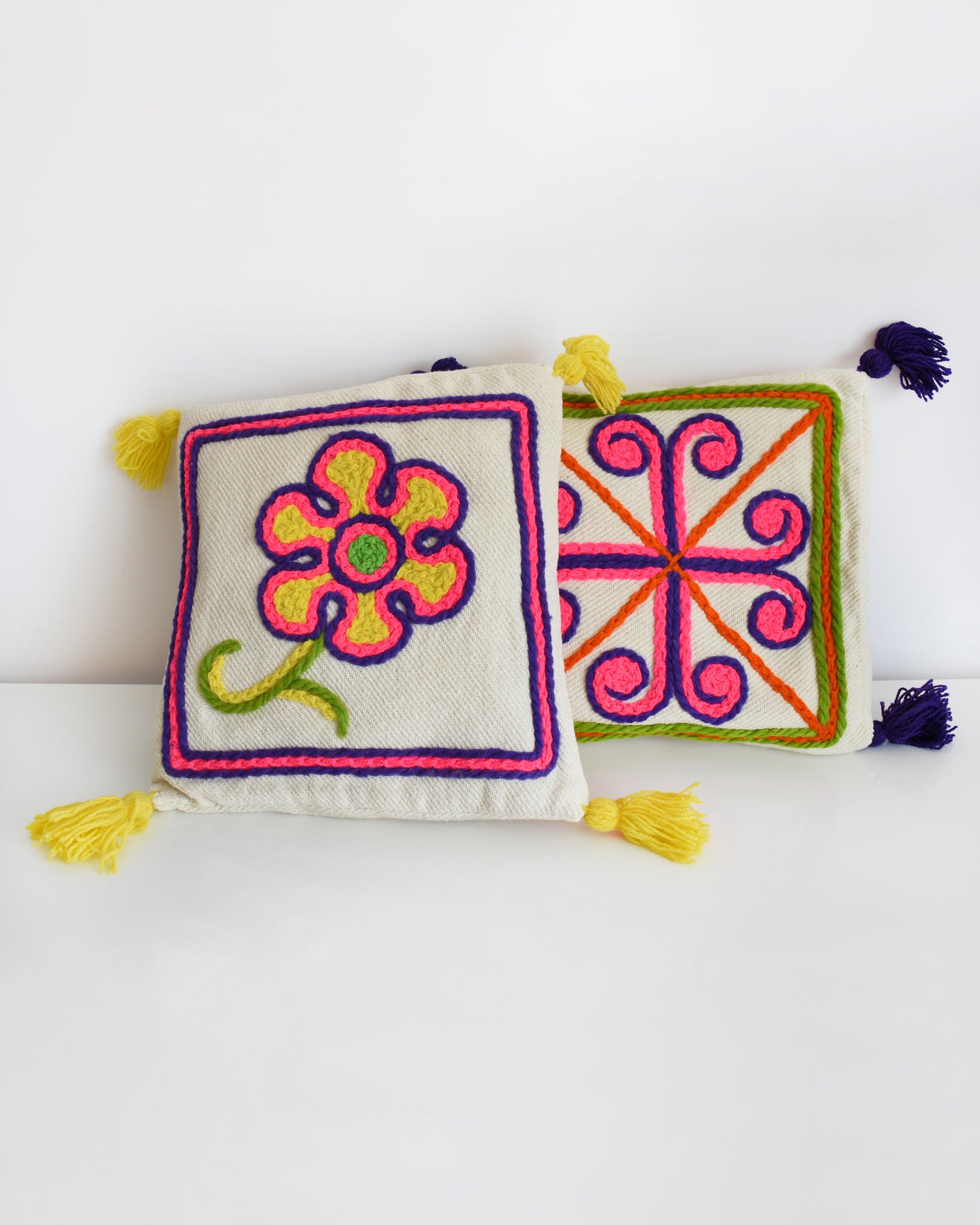 Two vintage crewel embroidered pillows on a table. One has a flower with yellow tassels and another has a curly cross with purple tassels.