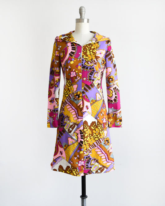 A vintage 1970s mod hooded dress features a vibrant geometric and floral print in shades of purple, pink, brown, yellow, and black. Half zipper on the front with long sleeves. The dress is on a dress form.
