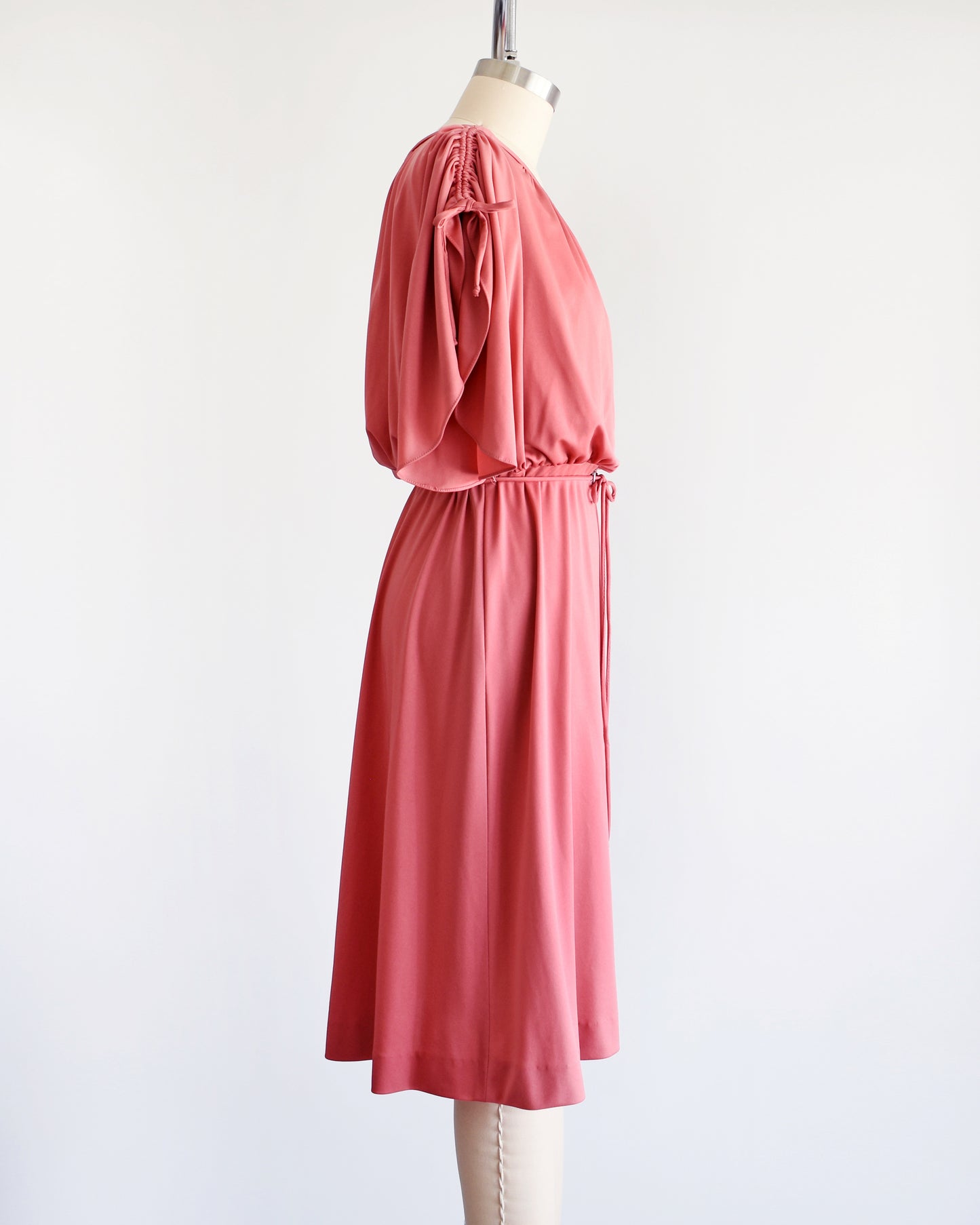 Side view of a vintage 1970s dusty pink dress with a blouson bodice and slit flutter sleeves that drawstrings at the end so you can adjust the length. Matching tie belt around the waist with a silver accent in the middle of the waist.