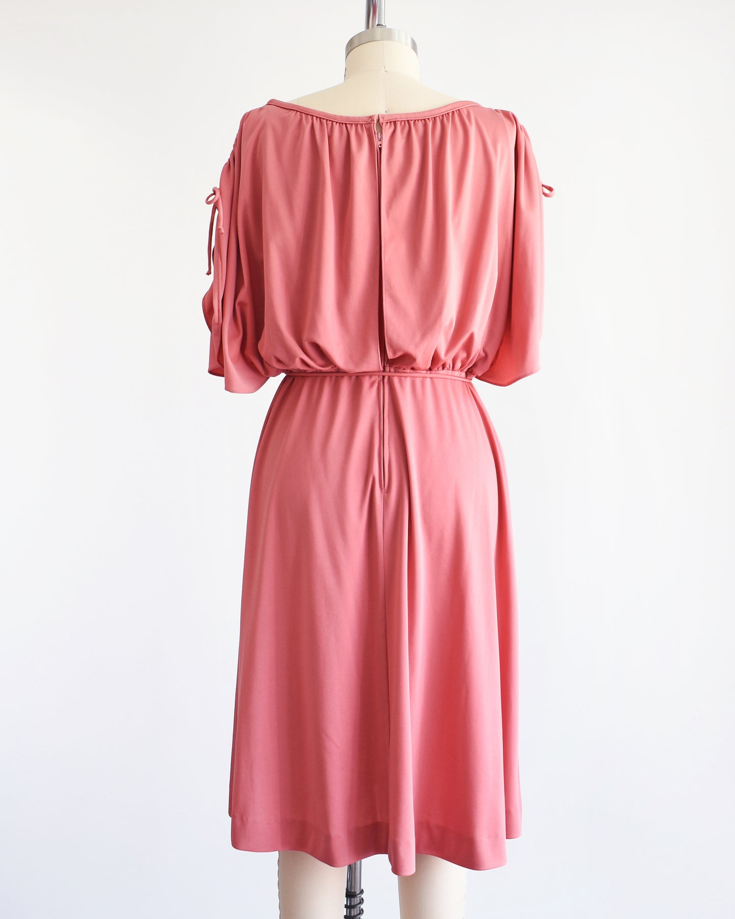 Back view of a vintage 1970s dusty pink dress with a blouson bodice and slit flutter sleeves that drawstrings at the end so you can adjust the length. Matching tie belt around the waist. Zipper up the back.