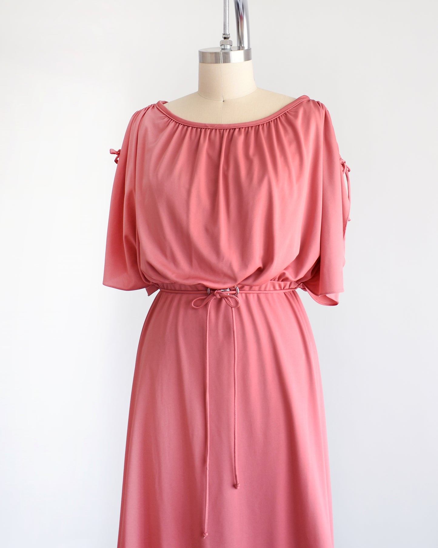 side front view of a vintage 1970s dusty pink dress with a blouson bodice and slit flutter sleeves that drawstrings at the end so you can adjust the length. Matching tie belt around the waist with a silver accent in the middle of the waist.