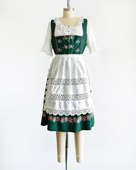 A vintage green dirndl dress that has pink and white embroidered flowers. The dress is worn with a white ruffled blouse and has a matching white apron. 