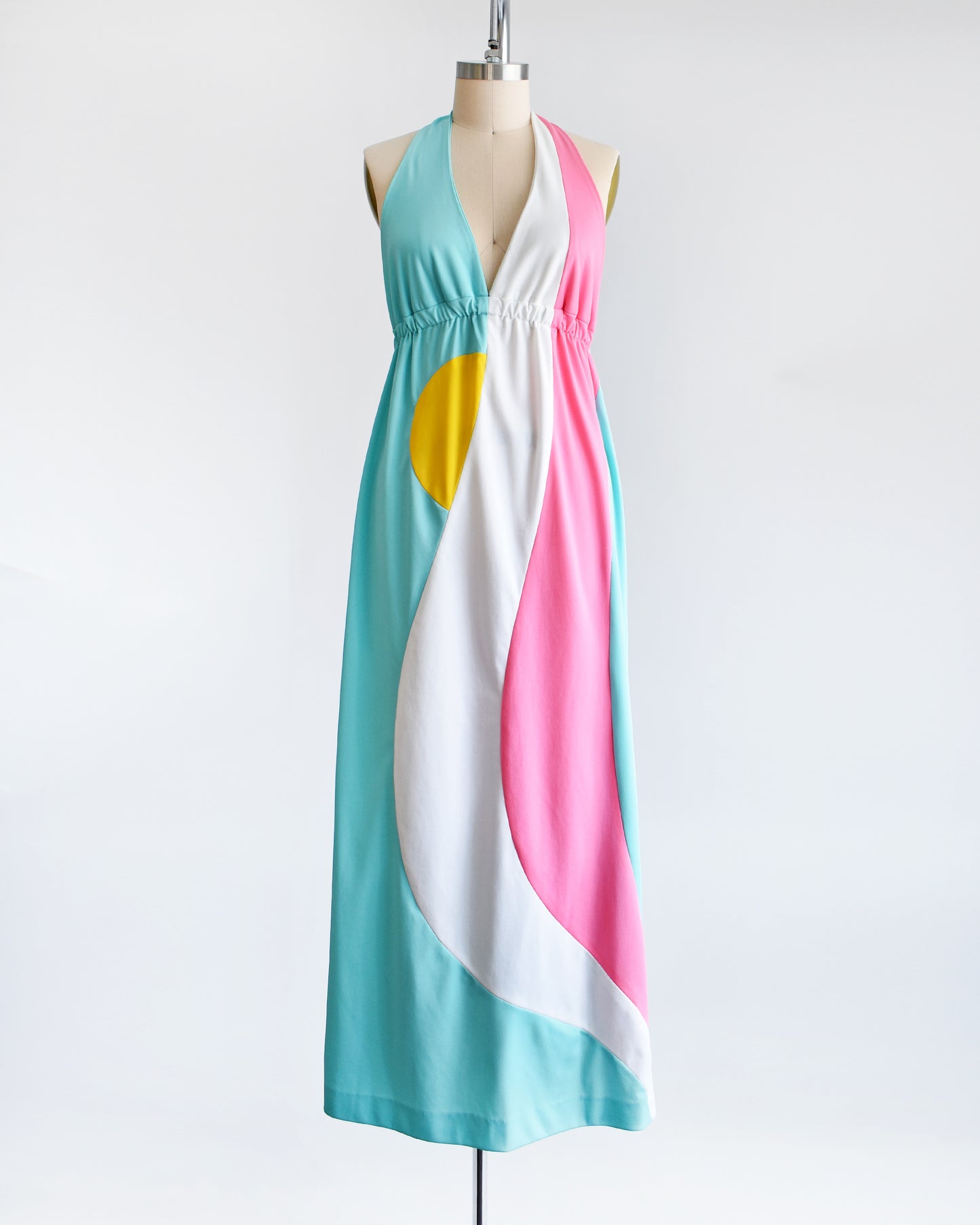 A vintage 1970s halter maxi dress that is blue with a white and pink swirl on the side with a half yellow circle peeking out just below the waistband.