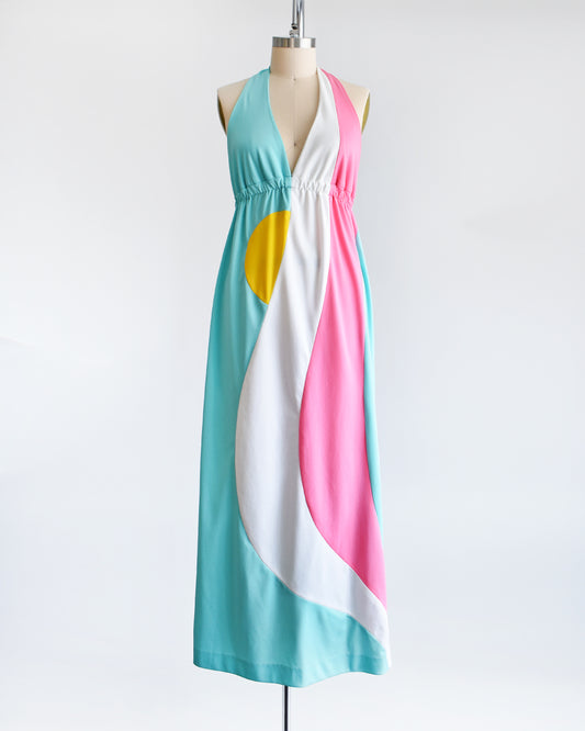 A vintage 1970s halter maxi dress that is blue with a white and pink swirl on the side with a half yellow circle peeking out just below the waistband.