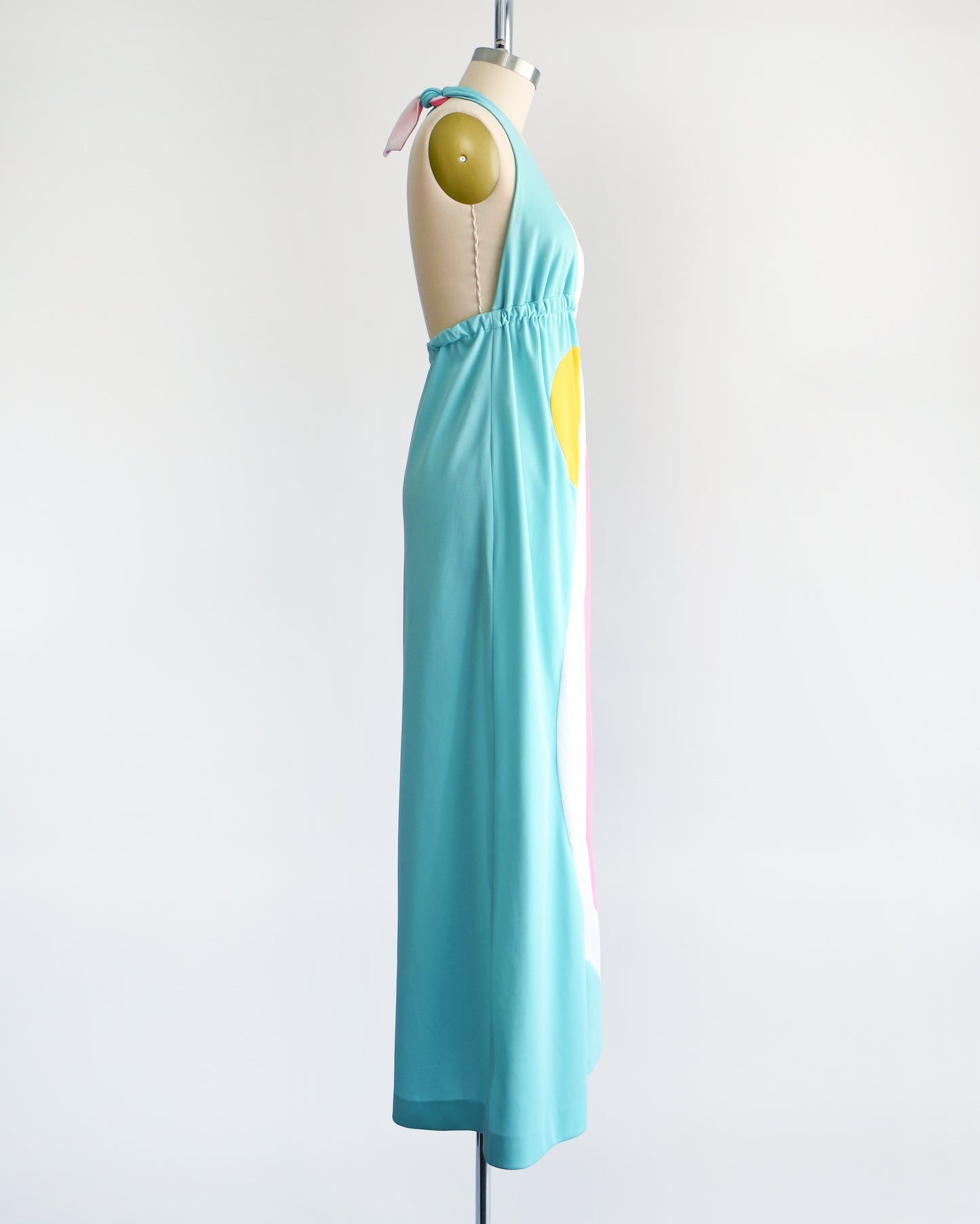Side view of a vintage 1970s halter maxi dress that is blue with a white and pink swirl on the side with a half yellow circle peeking out just below the waistband.