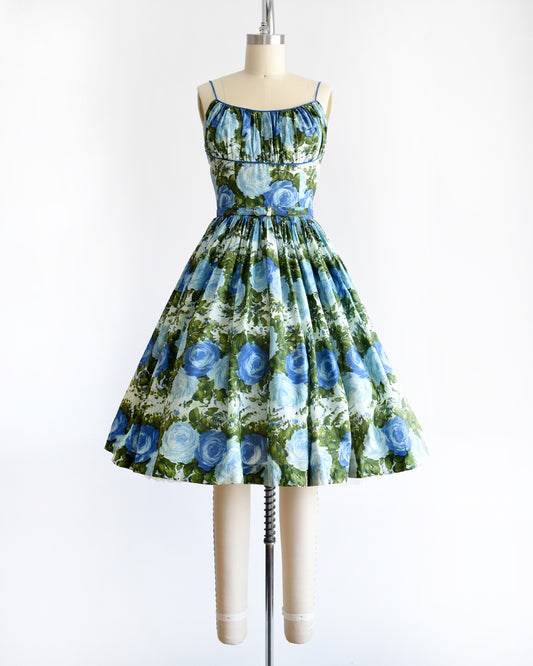 A gorgeous vintage 1950s cotton sundress features a beautiful dark and light blue floral print, with lush green leaves, set against a white background.