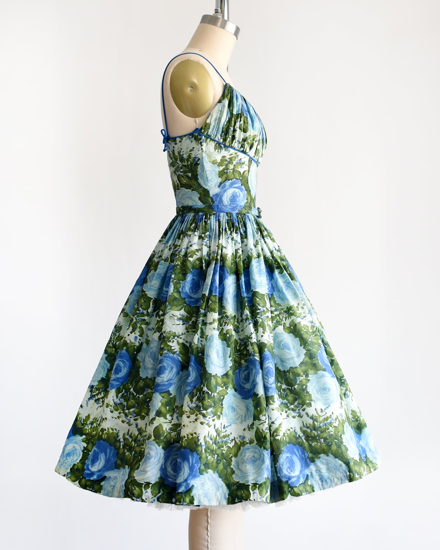 Side view of a vintage 1950s cotton sundress features a beautiful dark and light blue floral print, with lush green leaves, set against a white background.