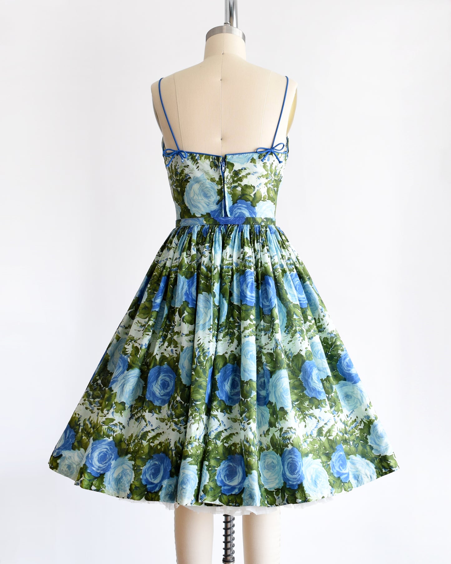 Back view of a vintage 1950s cotton sundress features a beautiful dark and light blue floral print, with lush green leaves, set against a white background. There is a metal zipper up the back and bows on the straps.