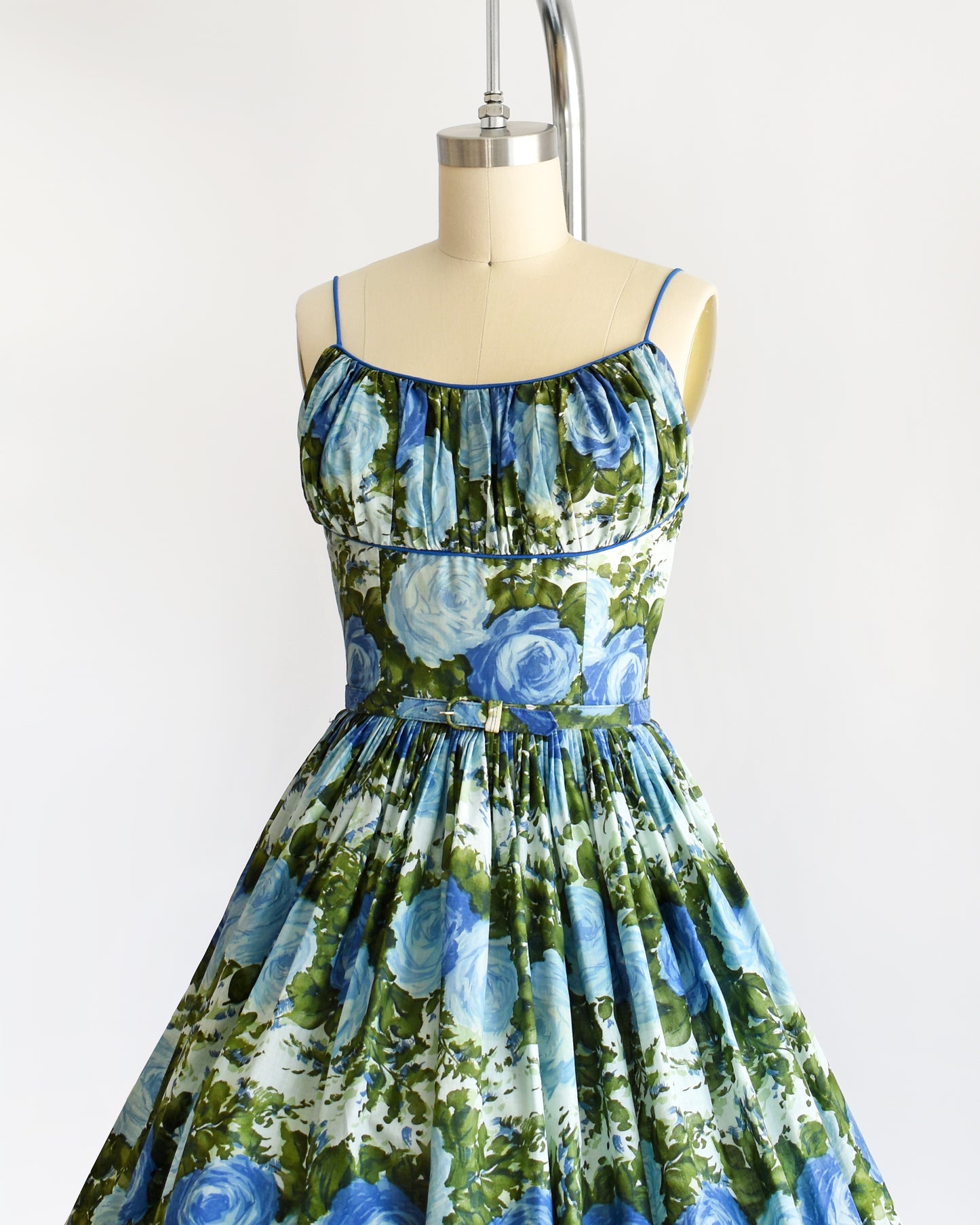 Side front view of a vintage 1950s cotton sundress features a beautiful dark and light blue floral print, with lush green leaves, set against a white background.