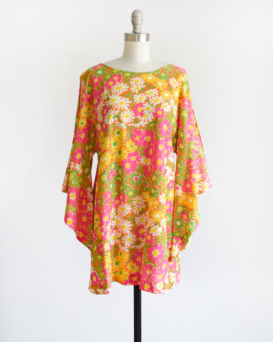 A vintage 1960s/1970s mini dress tunic that features a bold floral print in pink, yellow, white, and green against an orange backdrop with cascading angel sleeves.so the garment just slips on overhead.