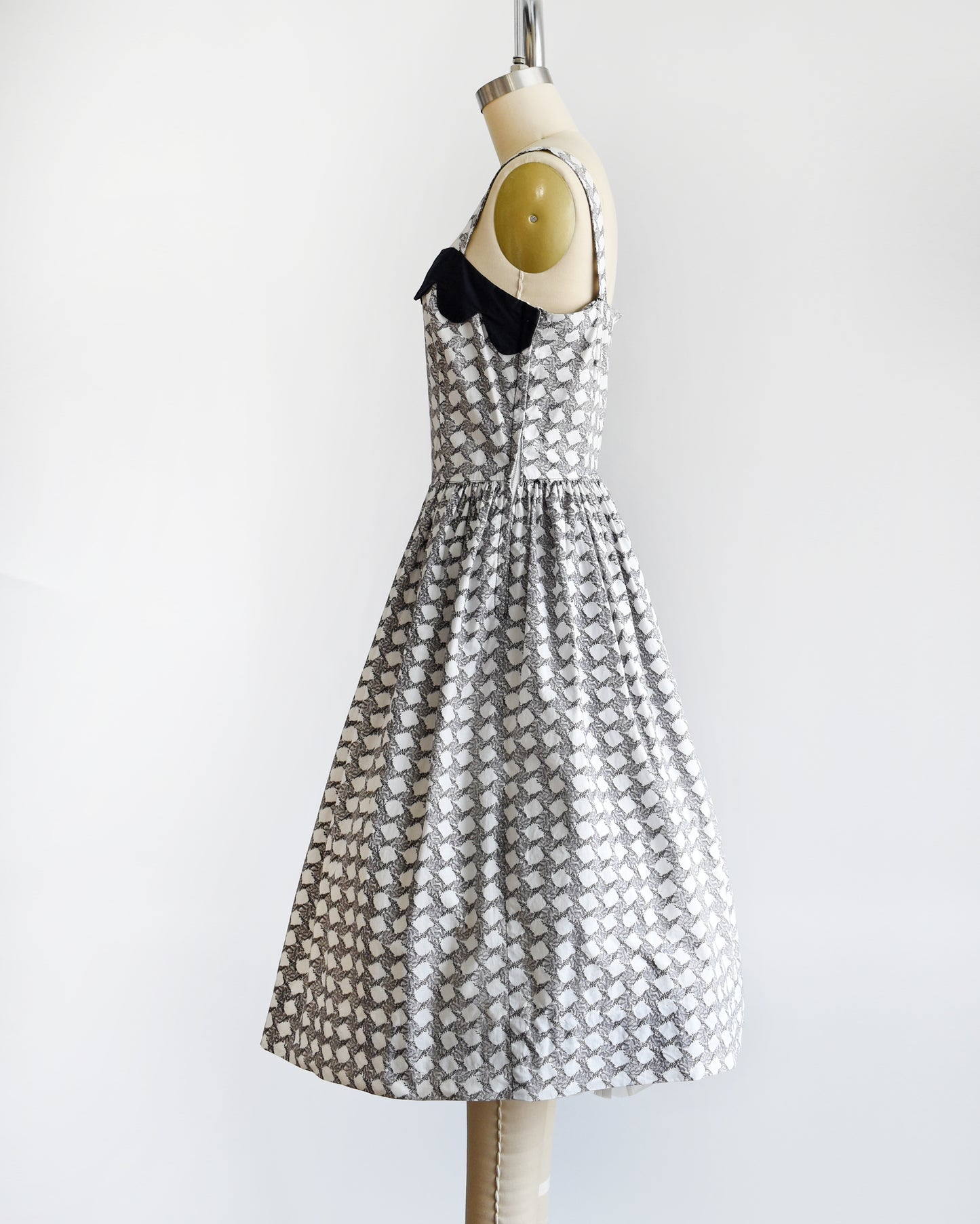 Side view of a vintage 1950s white dress with a  black fern motif that forms a diamond print. The dress has a black scalloped neckline and fit and flare skirt. A metal zipper is on the side.