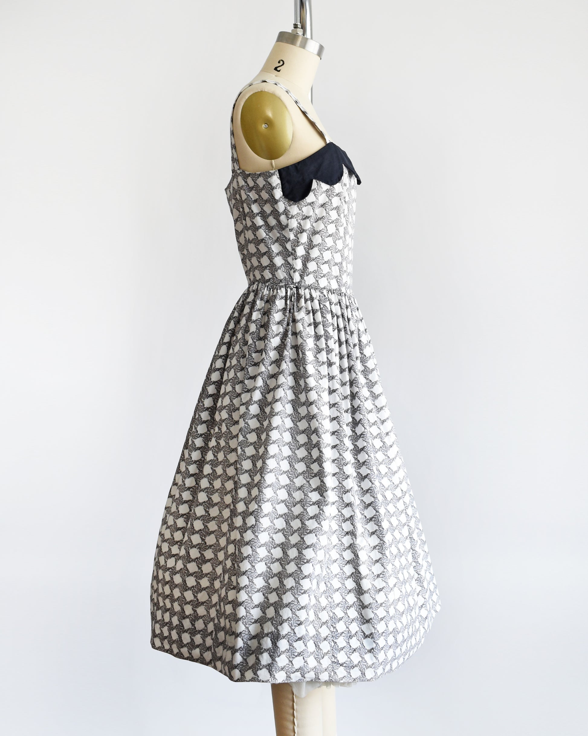 Side view of a vintage 1950s white dress with a  black fern motif that forms a diamond print. The dress has a black scalloped neckline and fit and flare skirt.
