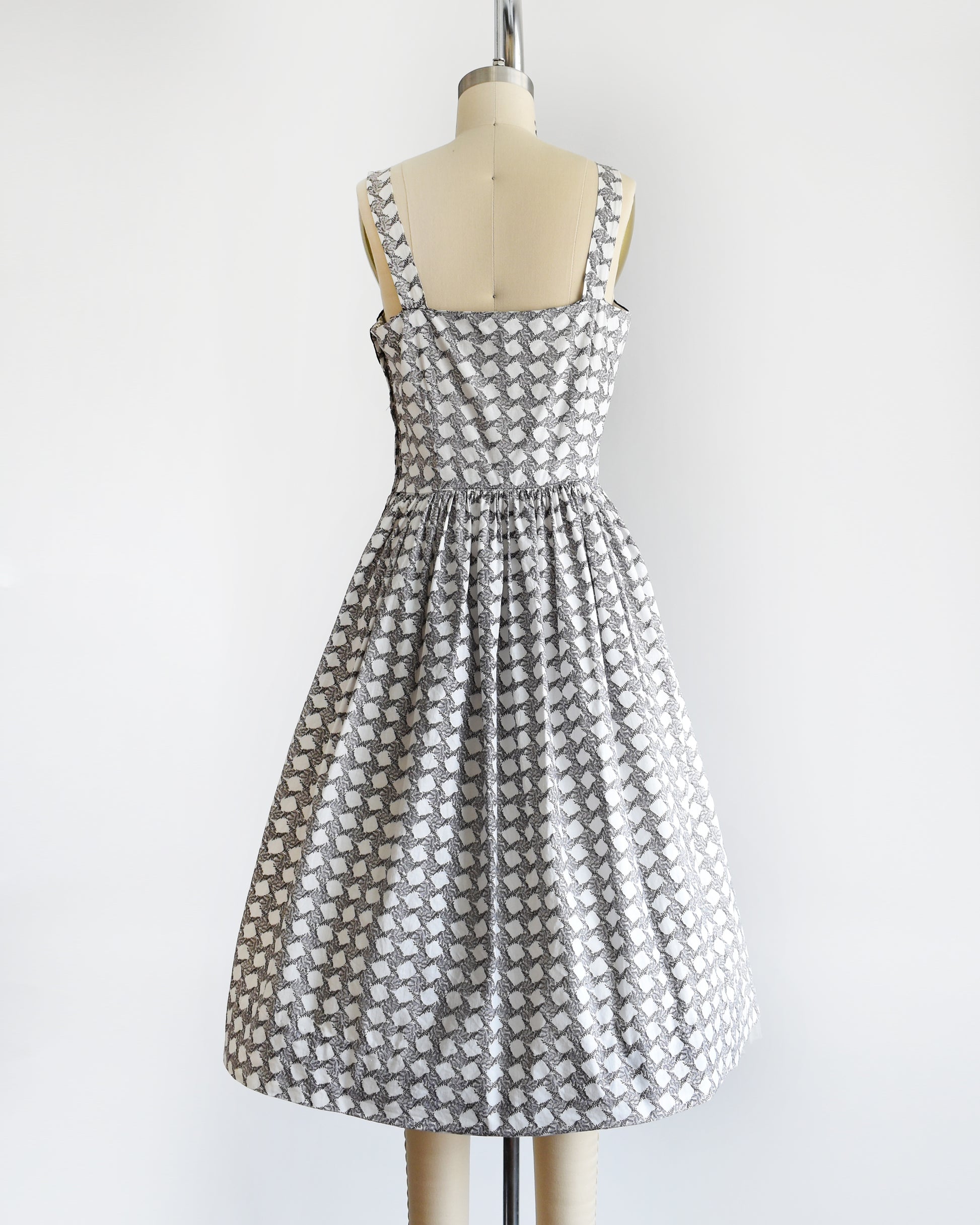 Back view of a vintage 1950s white dress with a  black fern motif that forms a diamond print. The dress has a black scalloped neckline and fit and flare skirt.