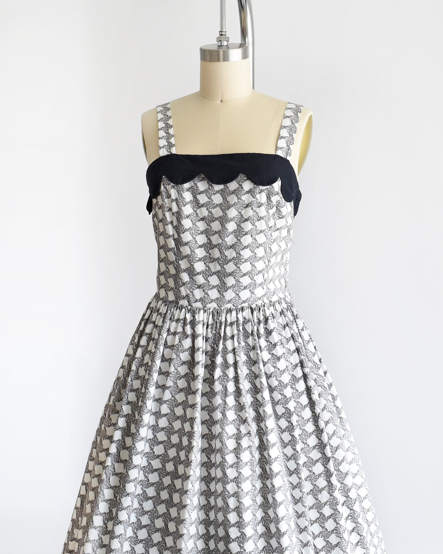 Side front view of a vintage 1950s white dress with a  black fern motif that forms a diamond print. The dress has a black scalloped neckline and fit and flare skirt.
