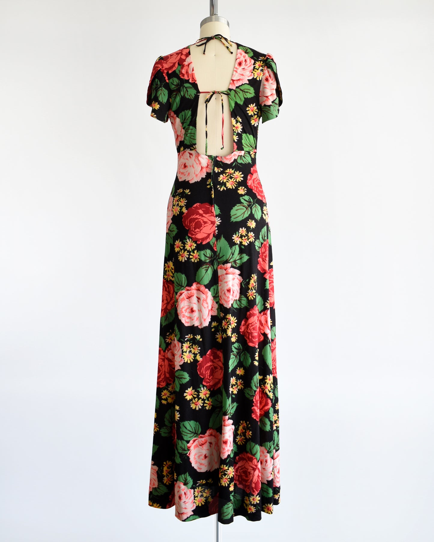 Back view of a A vintage 1970s black floral maxi dress features a vibrant floral pattern of red and pink roses, green leaves, and yellow and orange daisies. The upper part of the back is open and has two tie closures.