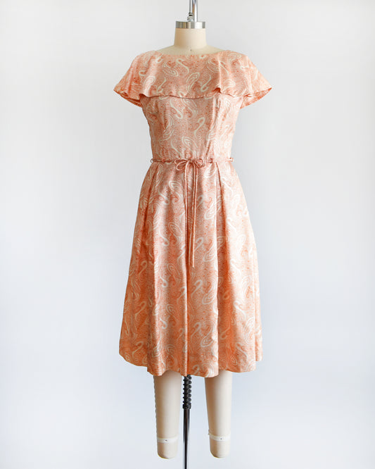 A vintage 1960s dress that features an orange and white paisley print, a ruffled collar, matching tie belt, and pleated skirt.