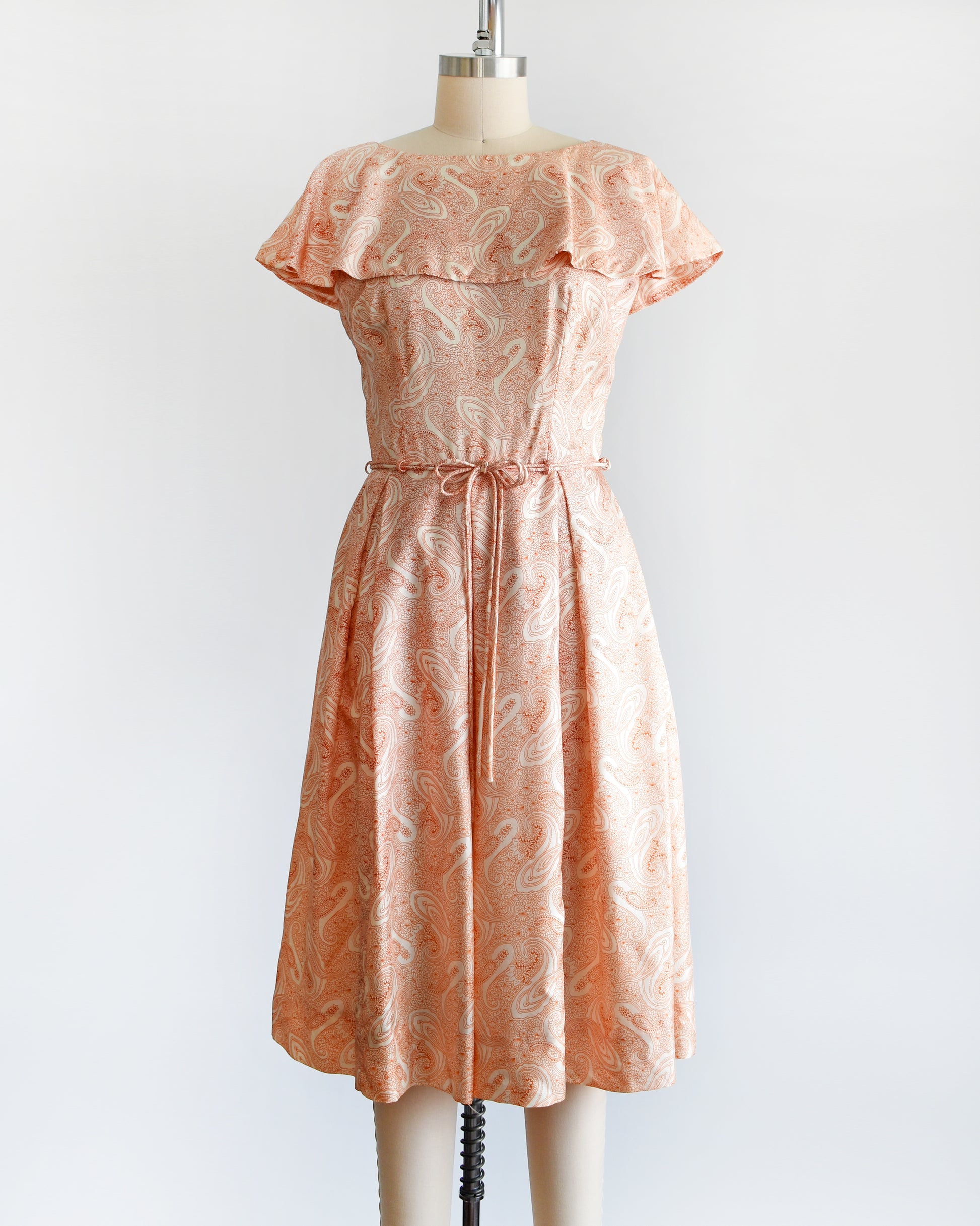 A vintage 1960s dress that features an orange and white paisley print, a ruffled collar, matching tie belt, and pleated skirt.