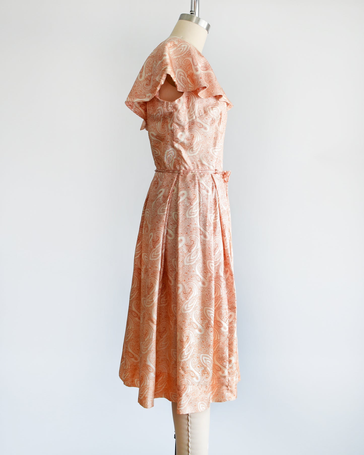 Side view of a vintage 1960s dress that features an orange and white paisley print, a ruffled collar, matching tie belt, and pleated skirt.