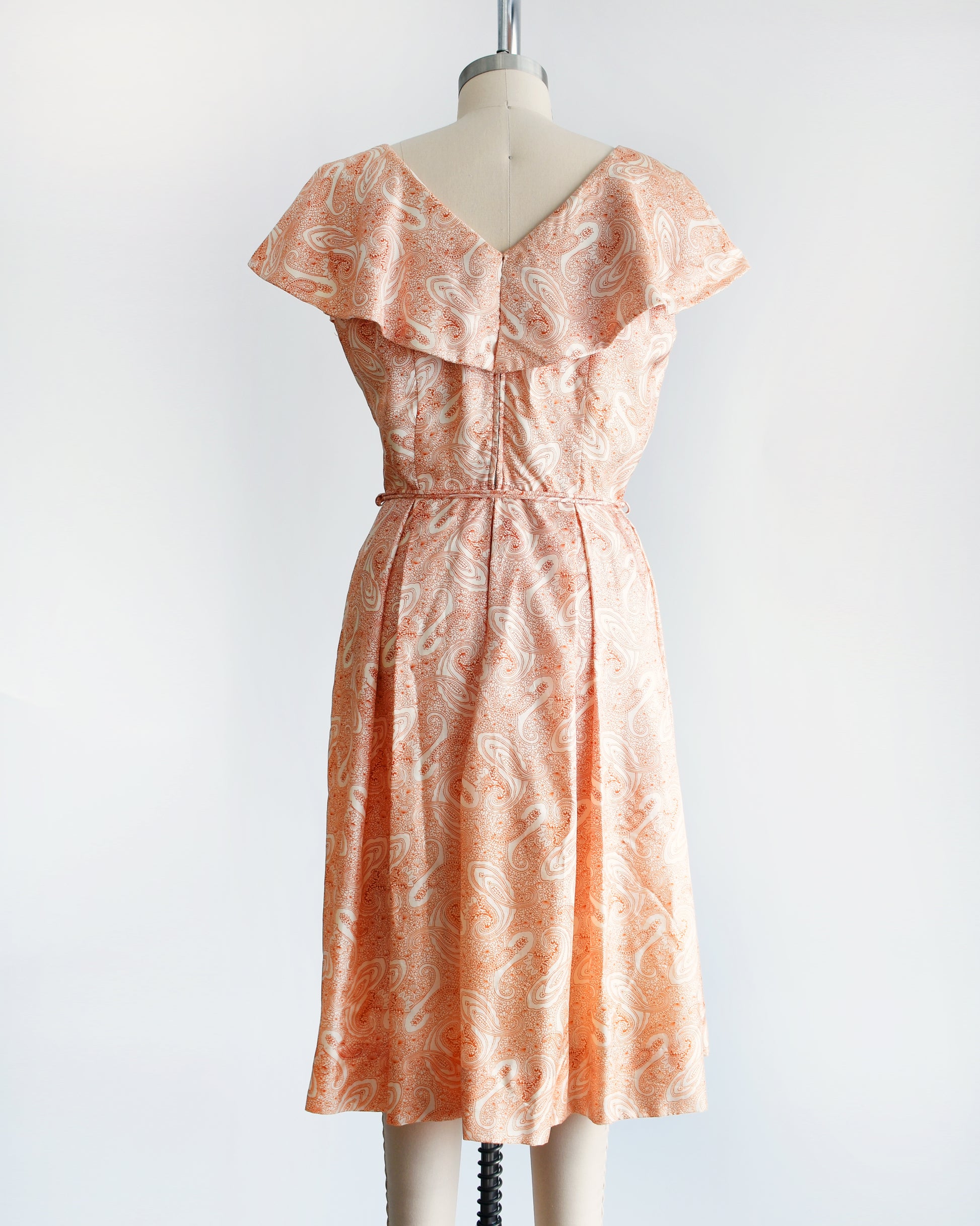 Back view of a vintage 1960s dress that features an orange and white paisley print, a ruffled neckline matching tie belt, and pleated skirt.