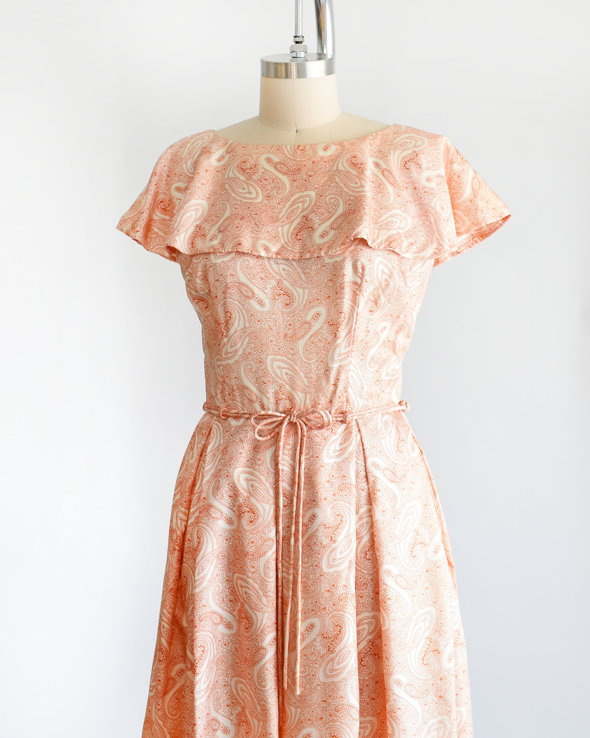 Side front view of a vintage 1960s dress that features an orange and white paisley print, a ruffled neckline, matching tie belt, and pleated skirt.