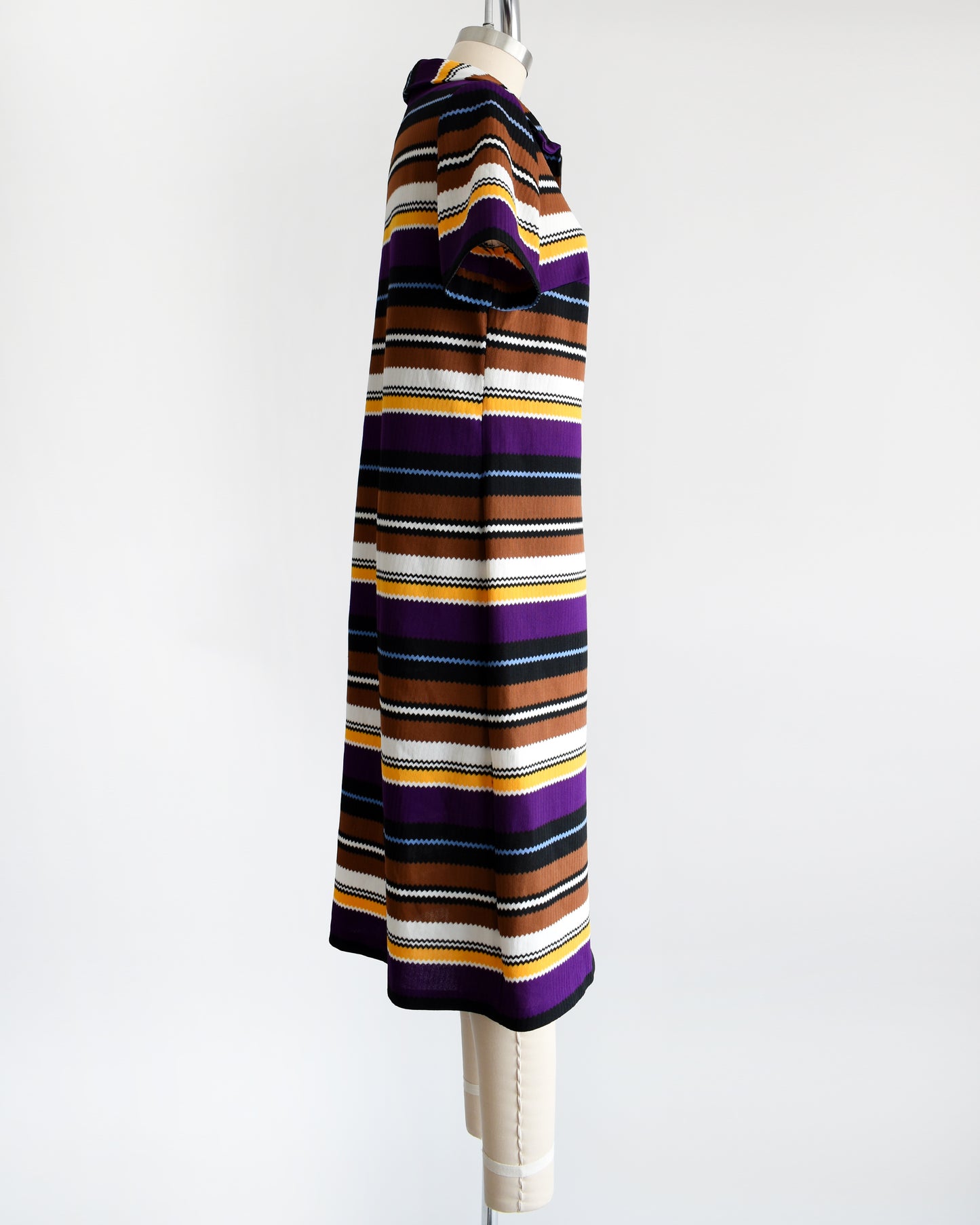 Side view of a vintage 1970s striped mod dress that has black, brown, white, blue, yellow, and purple horizontal stripes with zigzag edges.
