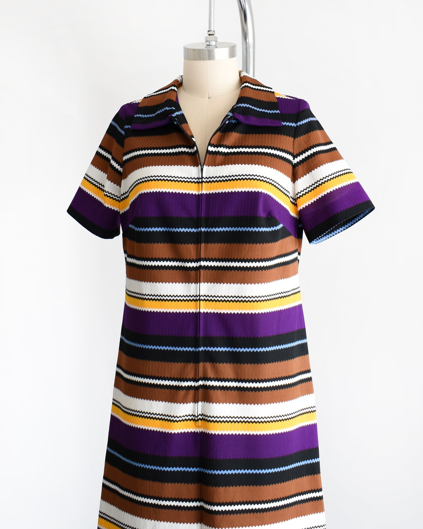 Side front view of a vintage 1970s striped mod dress that has black, brown, white, blue, yellow, and purple horizontal stripes with zigzag edges. The dress features a zipper down the front which is unzipped a little bit in this photo.