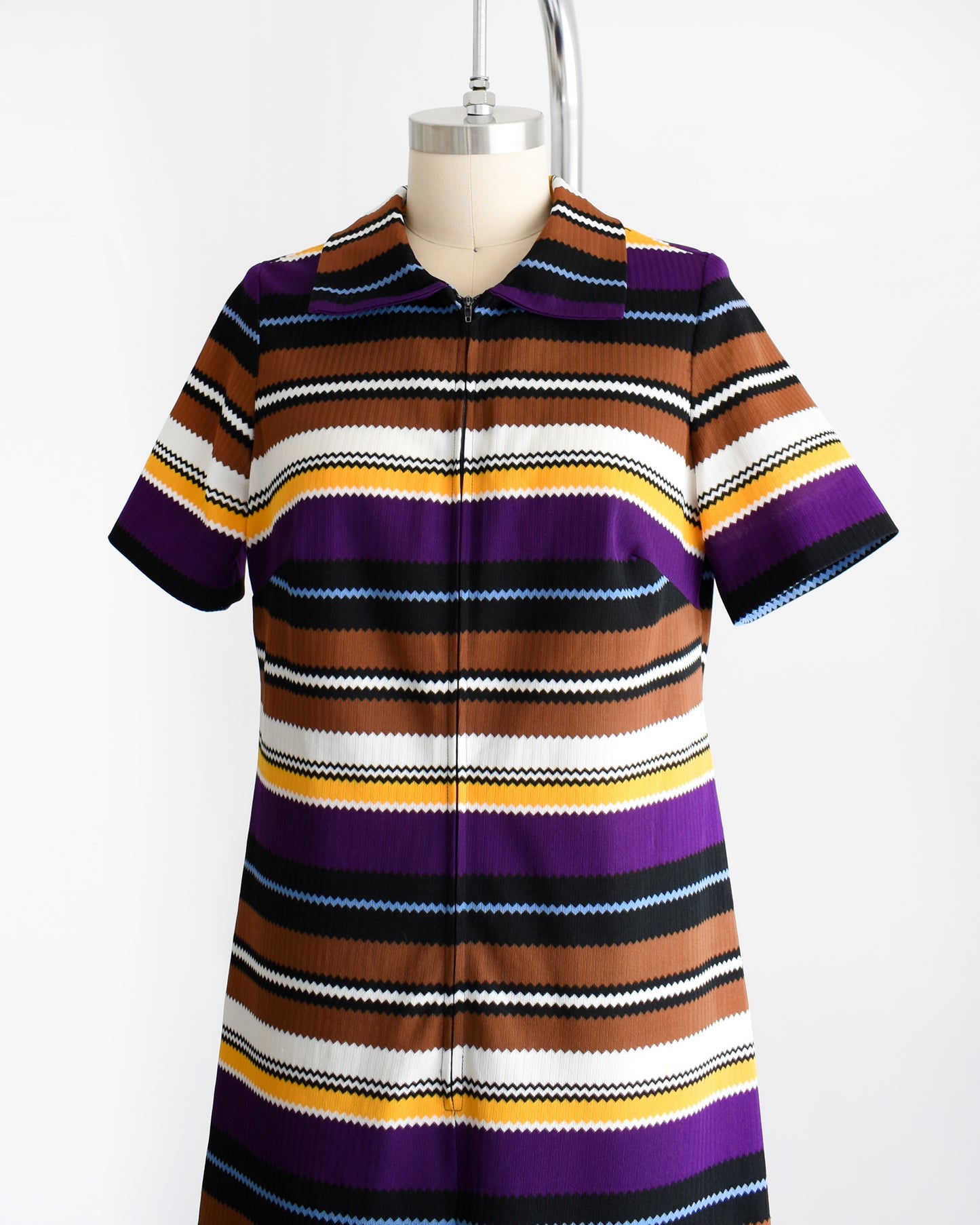Side front view of a vintage 1970s striped mod dress that has black, brown, white, blue, yellow, and purple horizontal stripes with zigzag edges. The dress features a zipper down the front which is zipped in this photo.