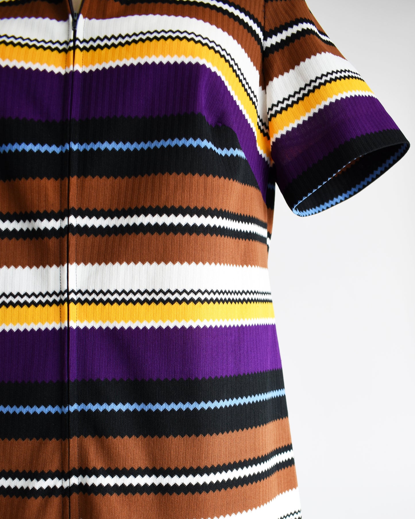 Close up of the a vintage 1970s striped mod dress that has black, brown, white, blue, yellow, and purple horizontal stripes with zigzag edges. The dress features a zipper down the front which is unzipped a little bit in this photo.