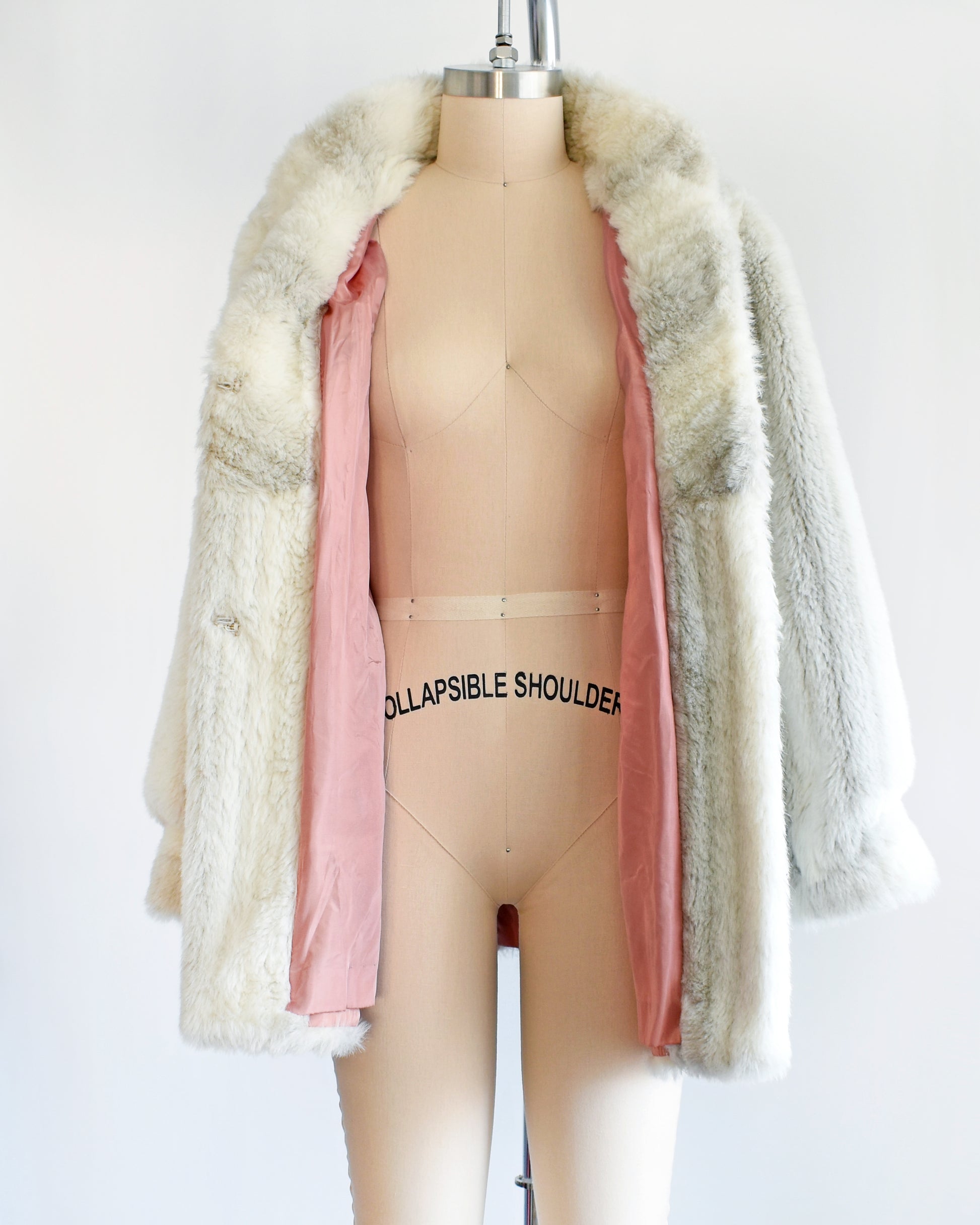 A vintage 70s/80s faux fur coat is made from a plush white faux fur with gray stripes, and features a stand-up collar with slightly puffed shoulders. The coat is opened revealing the salmon pink lining on the inside