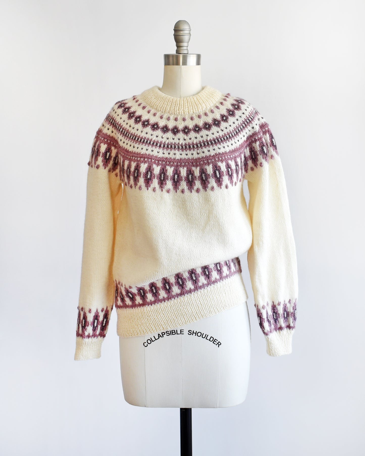 A vintage 1980s Dale of Norway cream wool sweater that has a purple Fair Isle pattern around the collar that's accented with matching purple nubby details. The sweater is slouched on one side