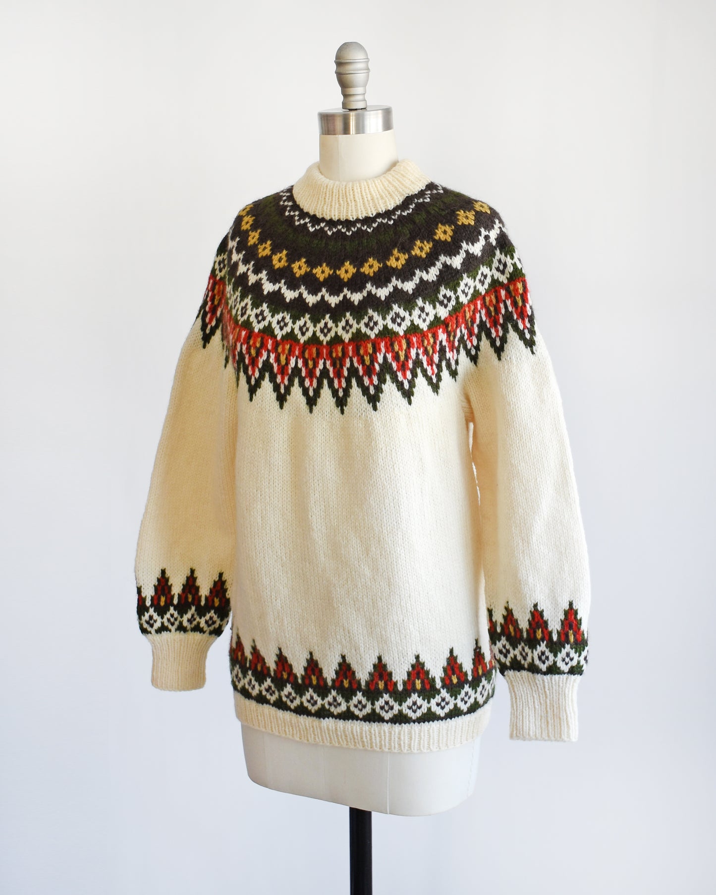Side front view of a vintage 1970s cream wool Nordic sweater with a brown, dark green, yellow, and red orange Fair Isle pattern around the collar and complimentary pattern on the hem and cuffs.