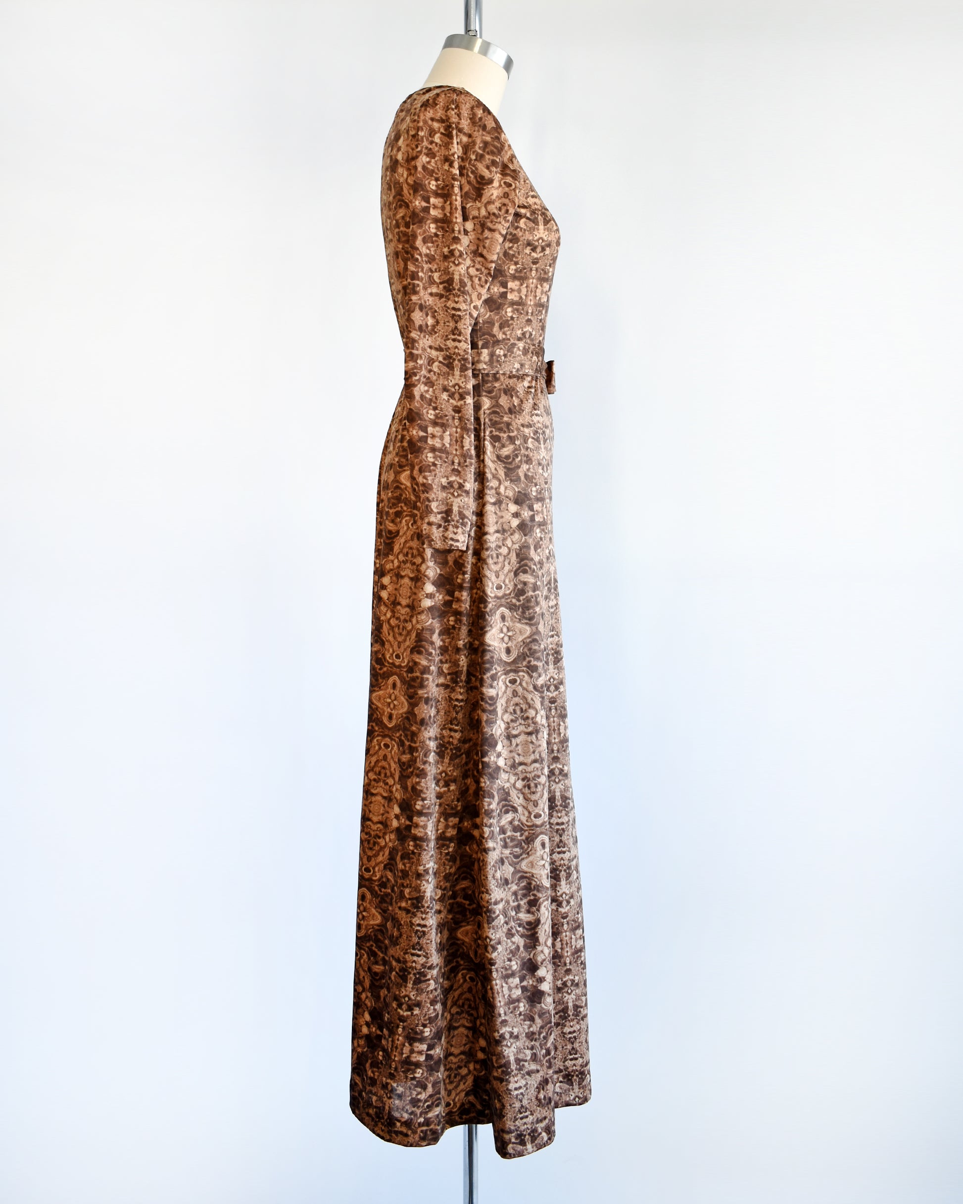 Side view of a vintage 1970s long sleeve maxi dress which features a psychedelic kaleidoscope pattern in light and dark brown.