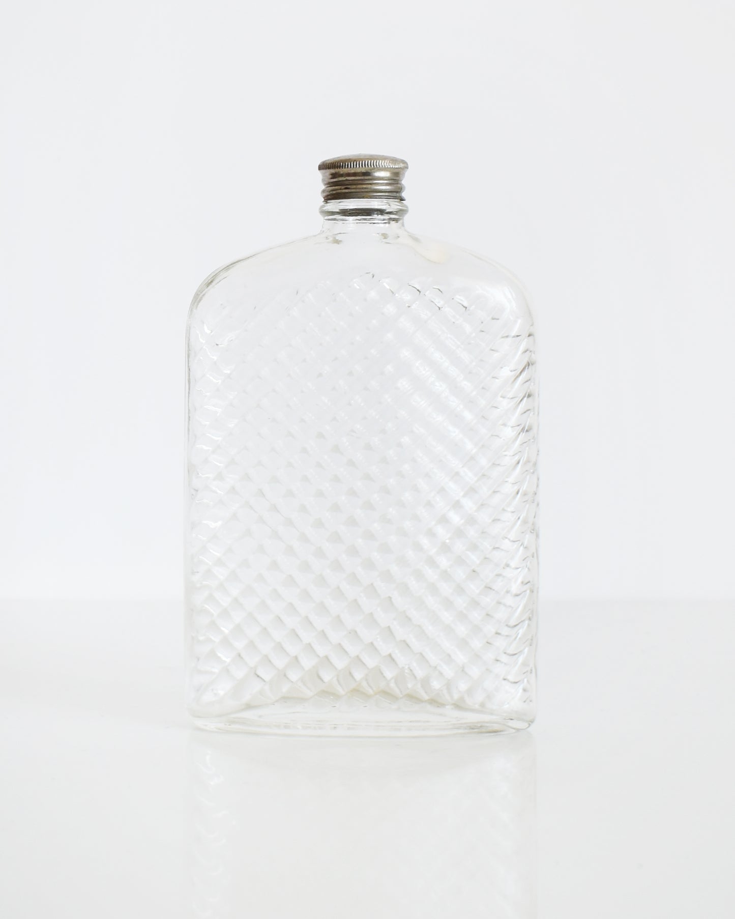 A vintage glass flask that has a cut diamond pattern and a silver screw top. The flask is on a white table and a white background.