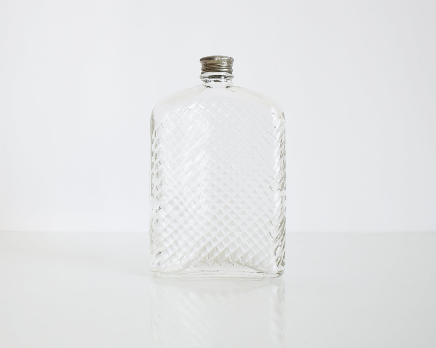 Back view of a vintage glass flask that has a cut diamond pattern and a silver screw top. The flask is on a white table and a white background.