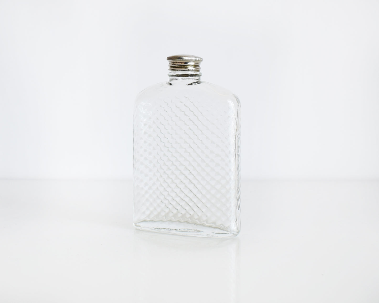 Side front view of a vintage glass flask that has a cut diamond pattern and a silver screw top. The flask is on a white table and a white background.